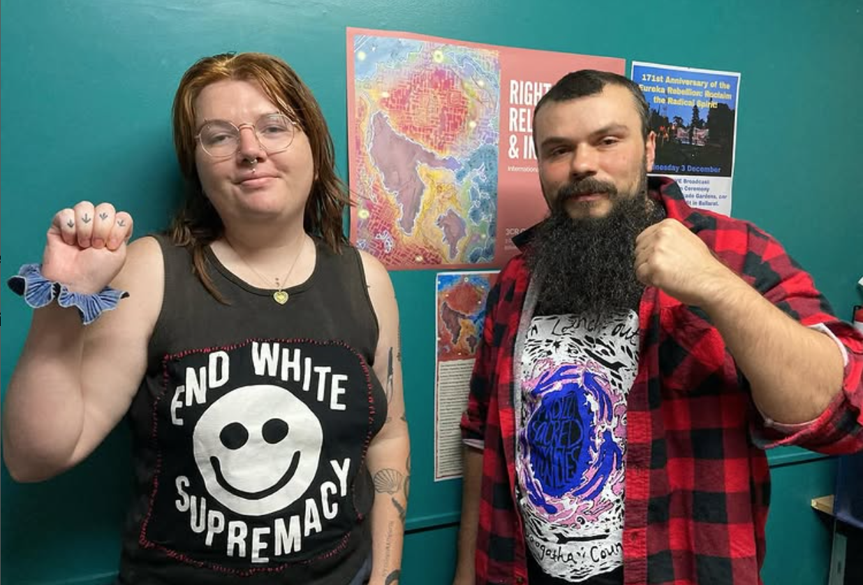 Image description: Em Randall, L, with light-coloured hair, wearing a t shirt that says ‘End White Supremacy’, standing next to Keiran Stewart-Assheton, R. They are in the 3CR studio with fists proudly raised in front of the disability day artwork by Renay Barker-Mulholland.