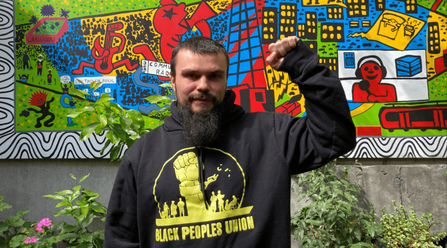 Keiran Stewart-Assheton in the 3CR garden wearing a Black Peoples Union hoodie and holding up a raised fist