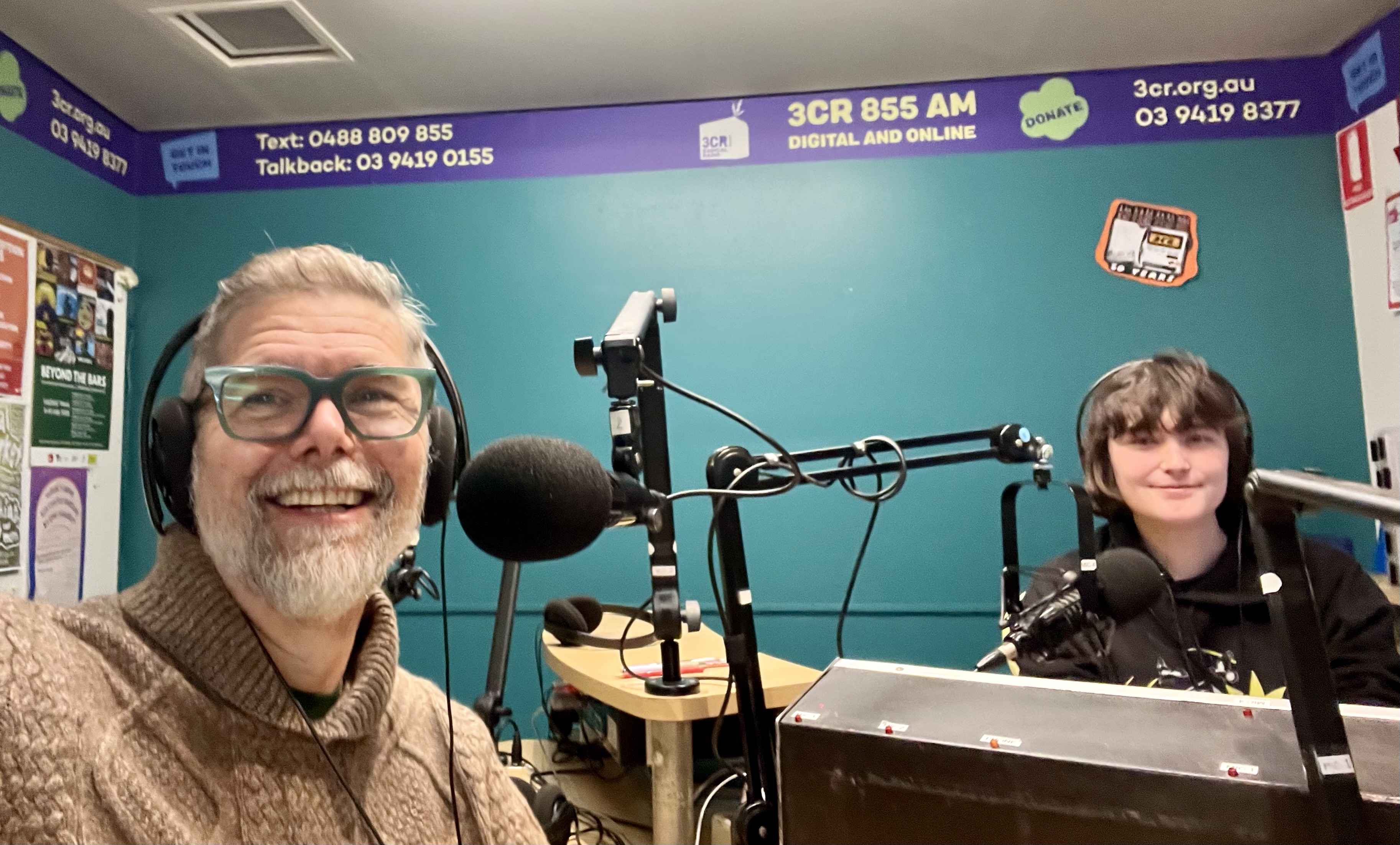 Amy and Keir sitting in the 3CR studio with their headphones on smiling for a sefie.