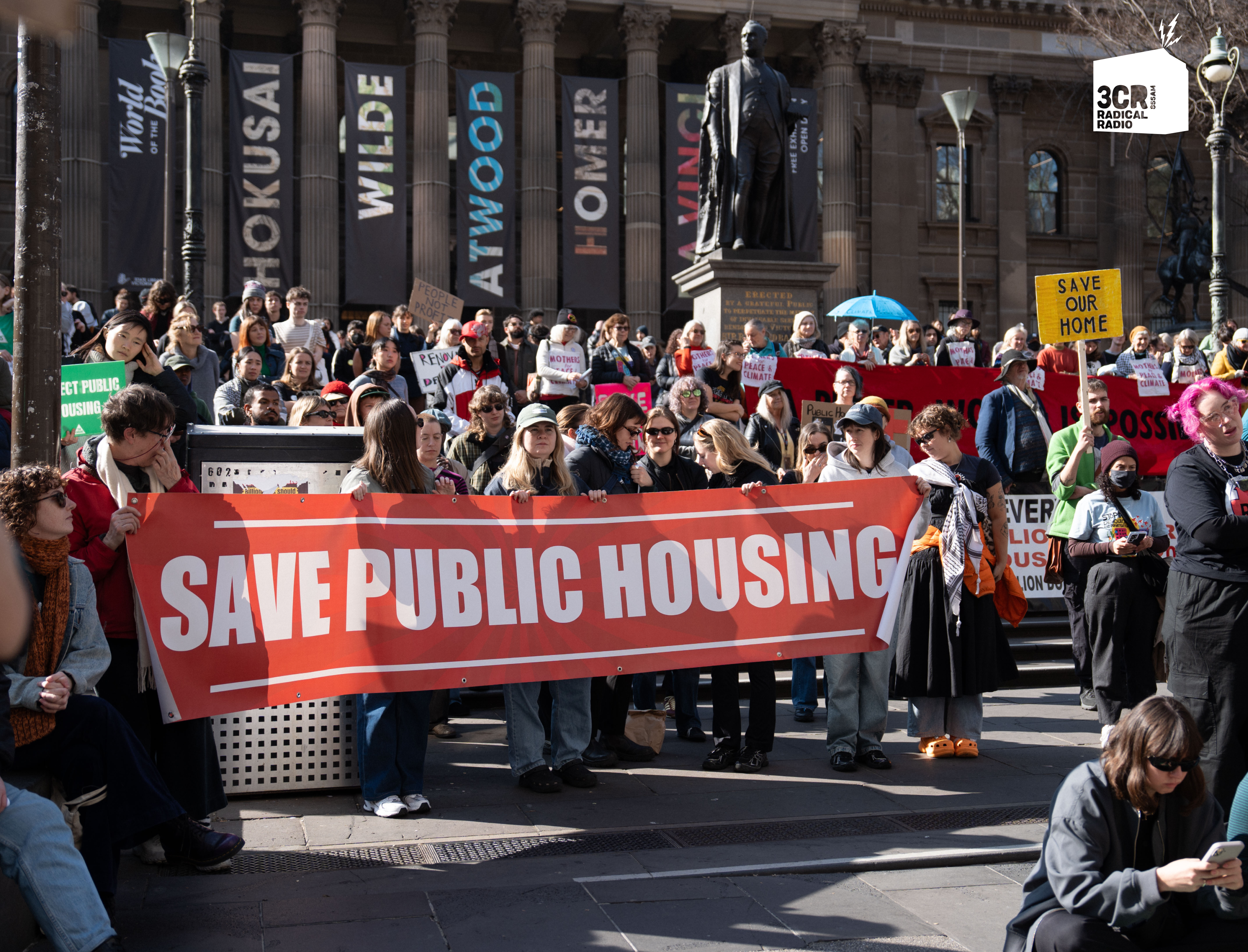 A group of people gathered at the Save Public Housing Rally hold a large sign reading “SAVE PUBLIC HOUSING.” Other smaller signs read “SAVE OUR HOME.” The crowd is standing in front of the State Library in Melbourne.