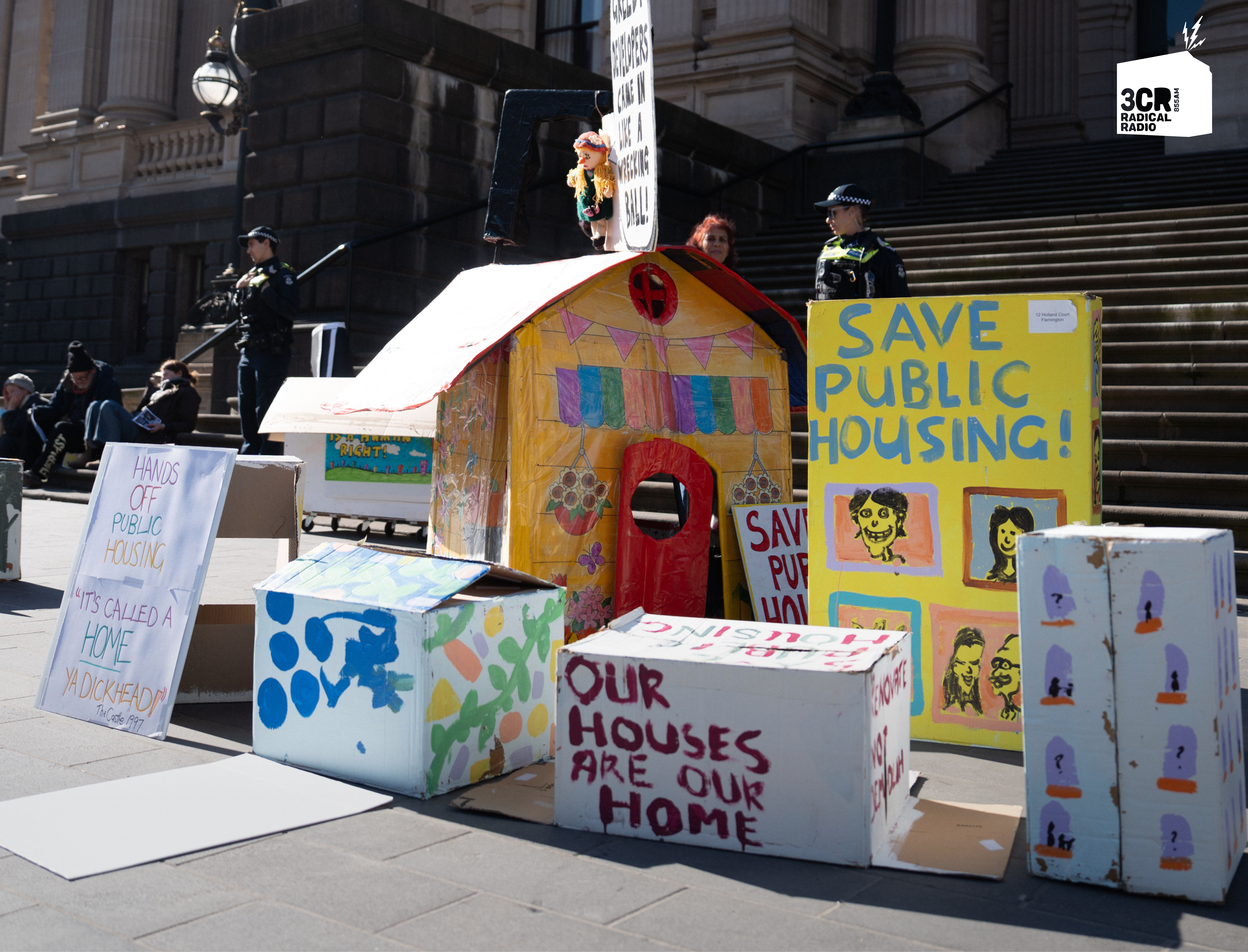 In front of the Parliament of Victoria, several cardboard houses display messages such as “SAVE PUBLIC HOUSING” and “OUR HOUSES ARE OUR HOME.” A police officer stands on the steps in the background.