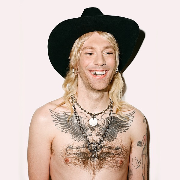 bare-chested person blonde hair black western hat tattoo on chest chains around neck
