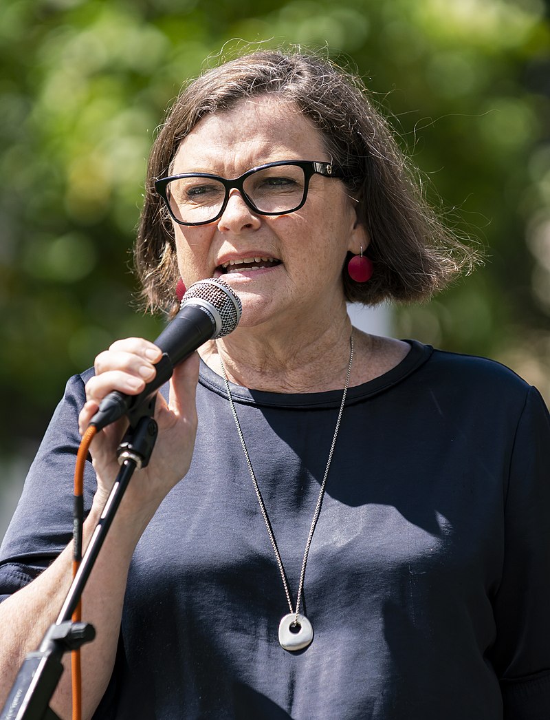 Woman with short dark hair black-rimmed glasses dark top holding microphone
