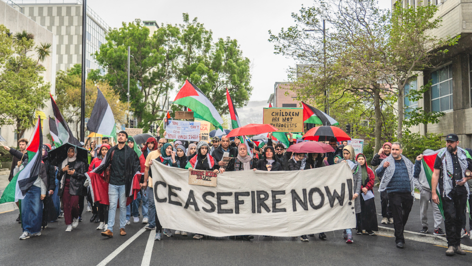 People marching in a pro-Palestine protest with a flag in front that reads 'Ceasfire now'
