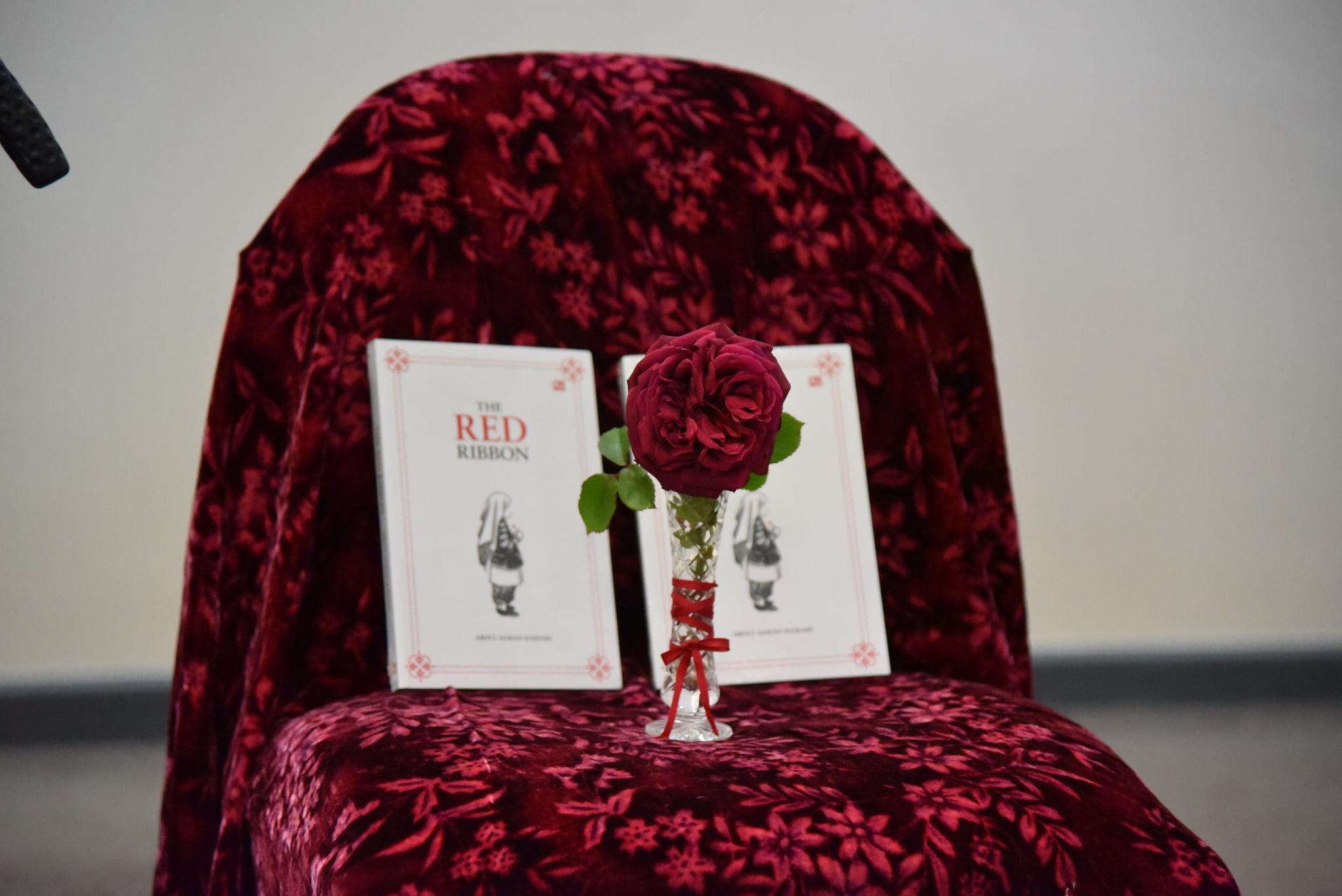 Red rose in a vase, resting on a chair, in front of two copies of the book, The Red Ribbon