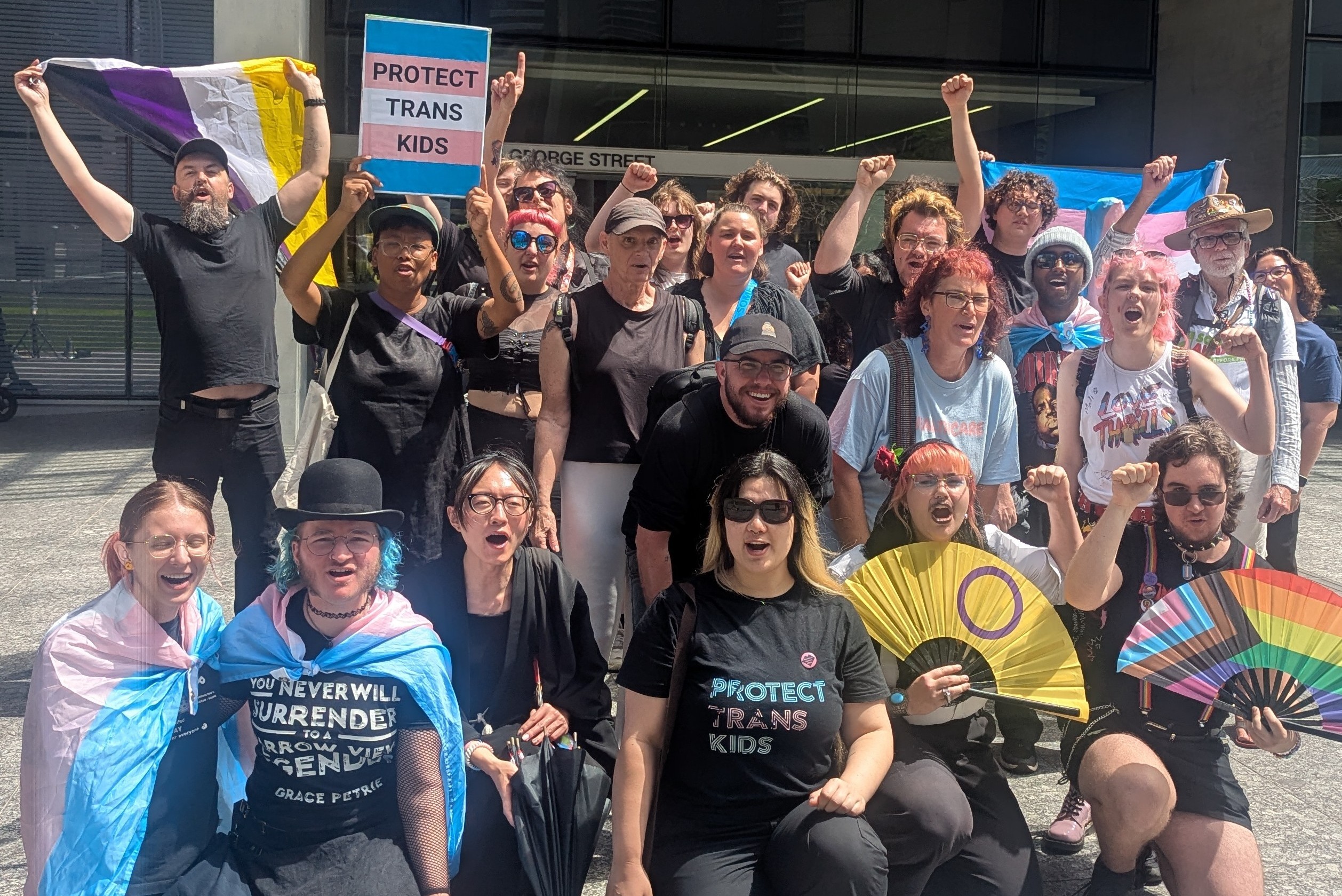 A group of trans rights activists pose and raise their fists and trans flags at a protest outside of the QLD Supreme Court