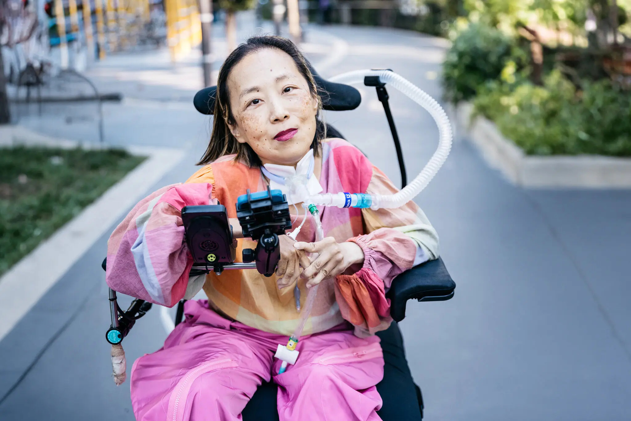 Image of Alice Wong, wearing a pink jumpsuit. She is sitting in a wheelchair with her mechanical ventilator. She is smiling at the camera. 