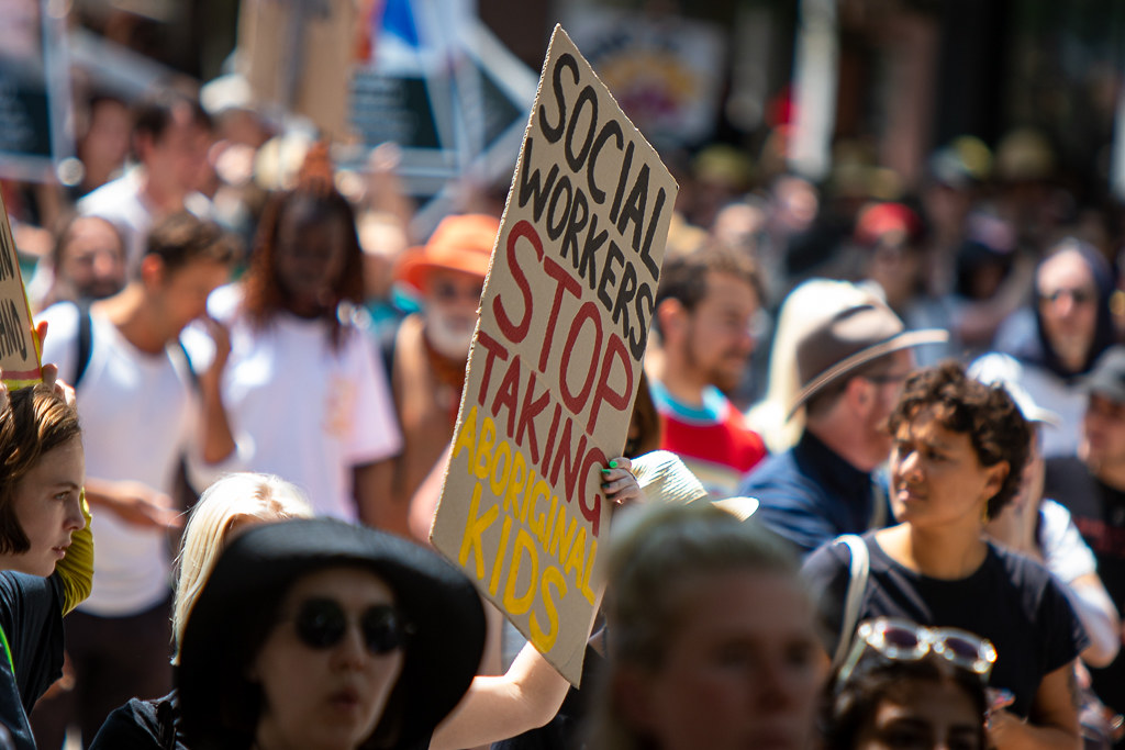 A photograph of a group of people at a protest, one of whom is holding up a painted cardboard sign which reads 'social workers stop taking Aboriginal kids' in black, red and yellow capital letters.