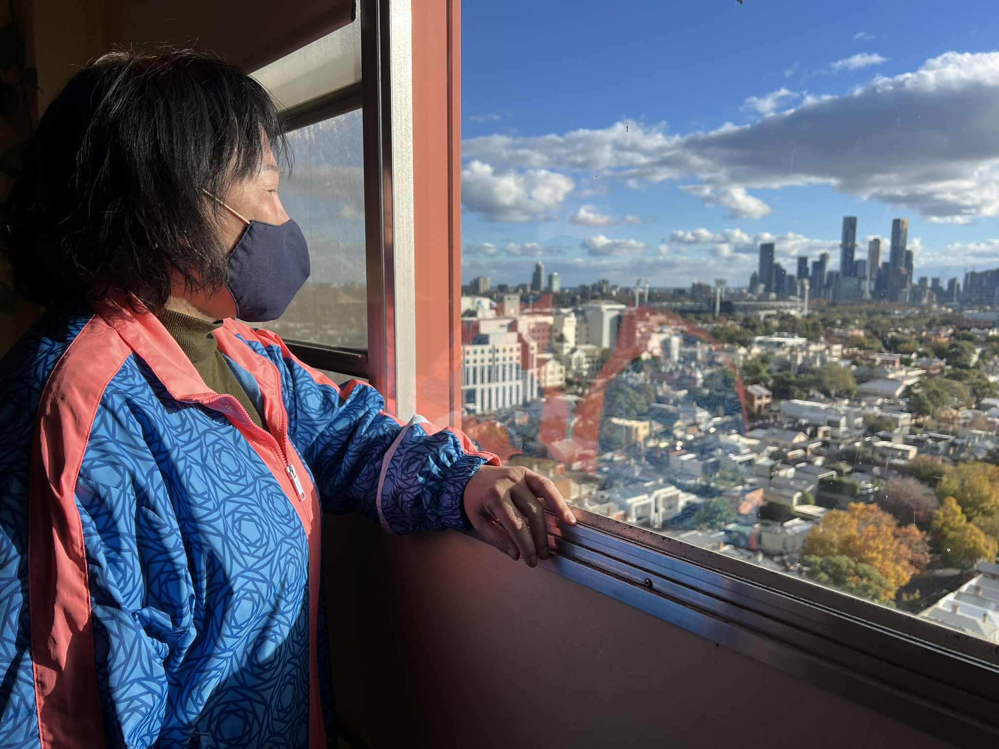 A Vietnamese resident of the housing estate looks out the window of her flat. You can see a view of Narrm. 