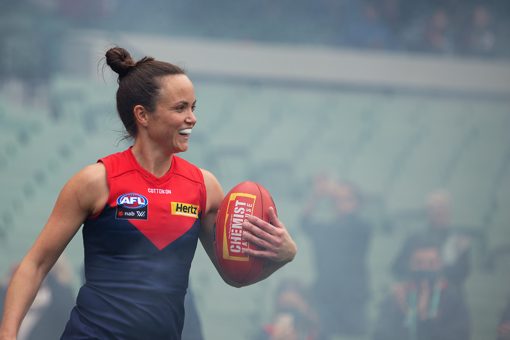 A close up photograph of Melbourne Demons AFLW captain Daisy Pearce standing in a stadium and holding a football in her left arm. She is smiling and wearing a Demons guernsey and her mouthguard. The background is hazy with some spectators visible.
