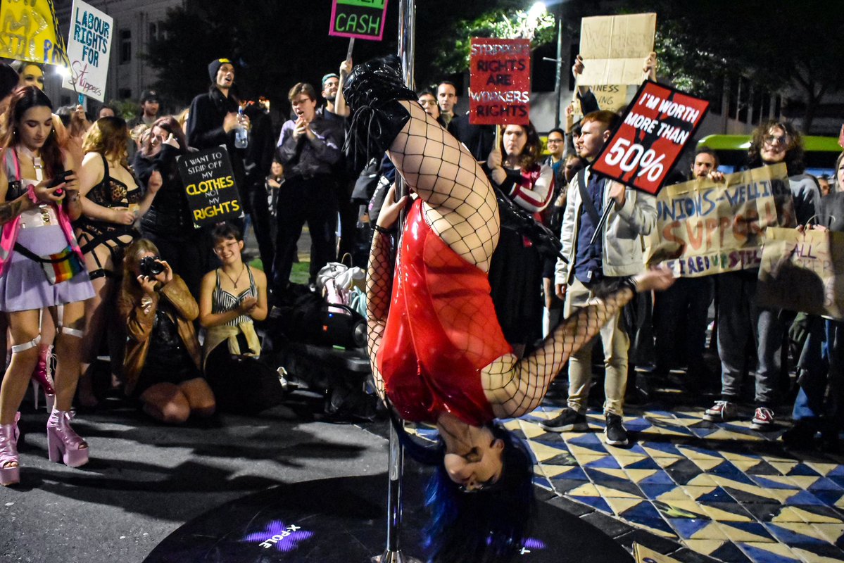 Image of a stripper dancing on a pole. The pole is out on the street and surrounding the stripper are a group of protestors protesting for stripper worker rights