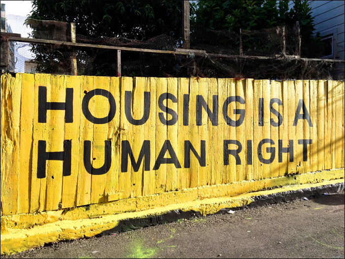 A high wooden fence painted bright yellow, with the words 'housing is a human right' painted over the top in large black capital letters.