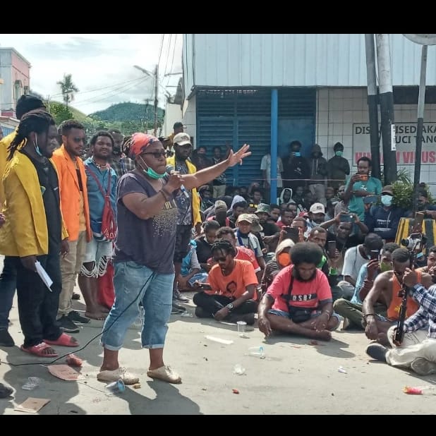 A photograph of West Papuan feminist Esther Haluk speaking at a peaceful demonstration against the break up of the provinces. She is standing to the left of the photograph, speaking into a microphone and gesturing to a crowd seated on the ground.