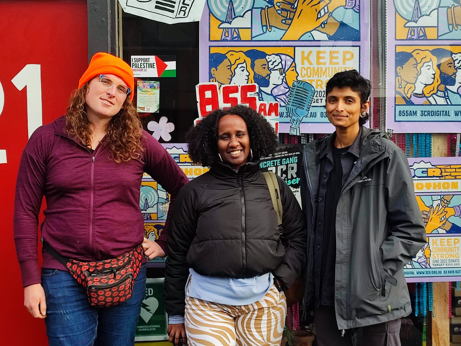 A photograph of Iris, Ayan and Priya standing in front of the 3CR station. The front window behind them is covered in 2022 Radiothon posters.