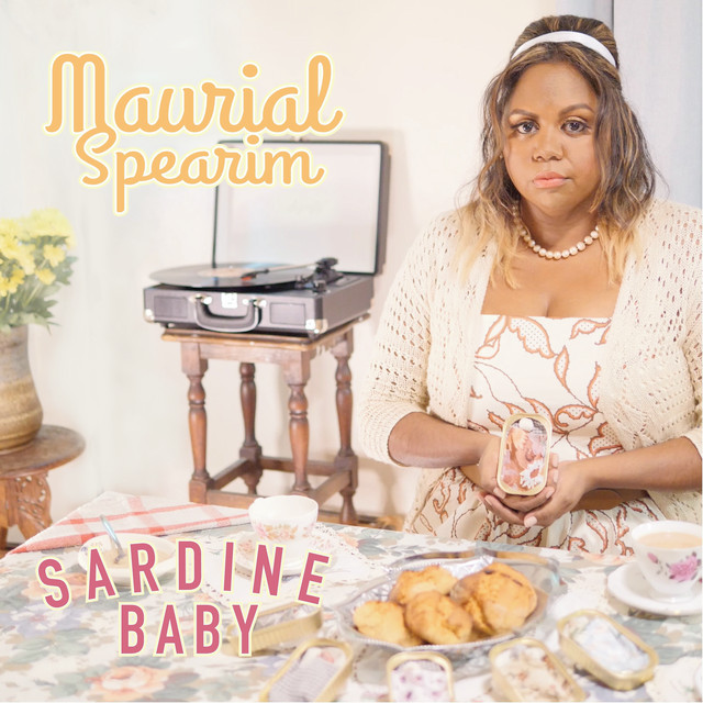 The cover for Maurial Spearim's single Sardine Baby. Maurial, dressed in 1950s femme style, sits at a table holding a Sardine Baby (tin with a pebble and cloth). The table is laid for tea, with a plate of scones, teacups, and more Sardine Babies. In the background is a record player on a small table.