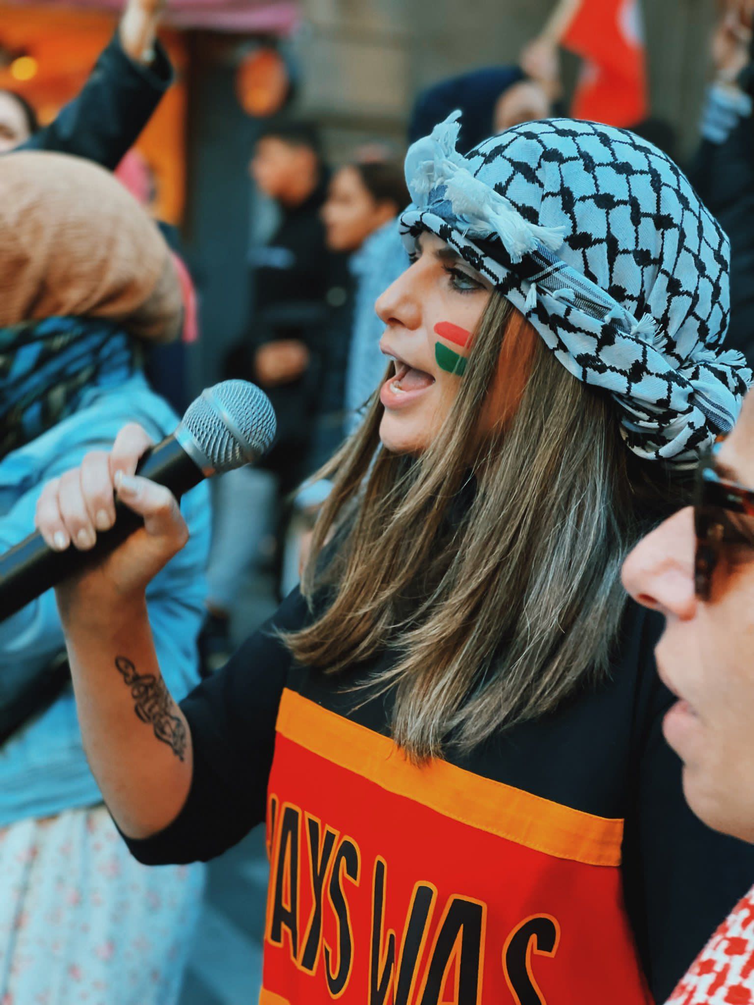 Palestinian activist, campaigner, and storyteller Jeanine Hourani speaks to a microphone at a protest. She is wearing a black and white kuffiyeh (كُوفِيَّة‎) with a black, yellow and red Always Was (Aboriginal land) sweater