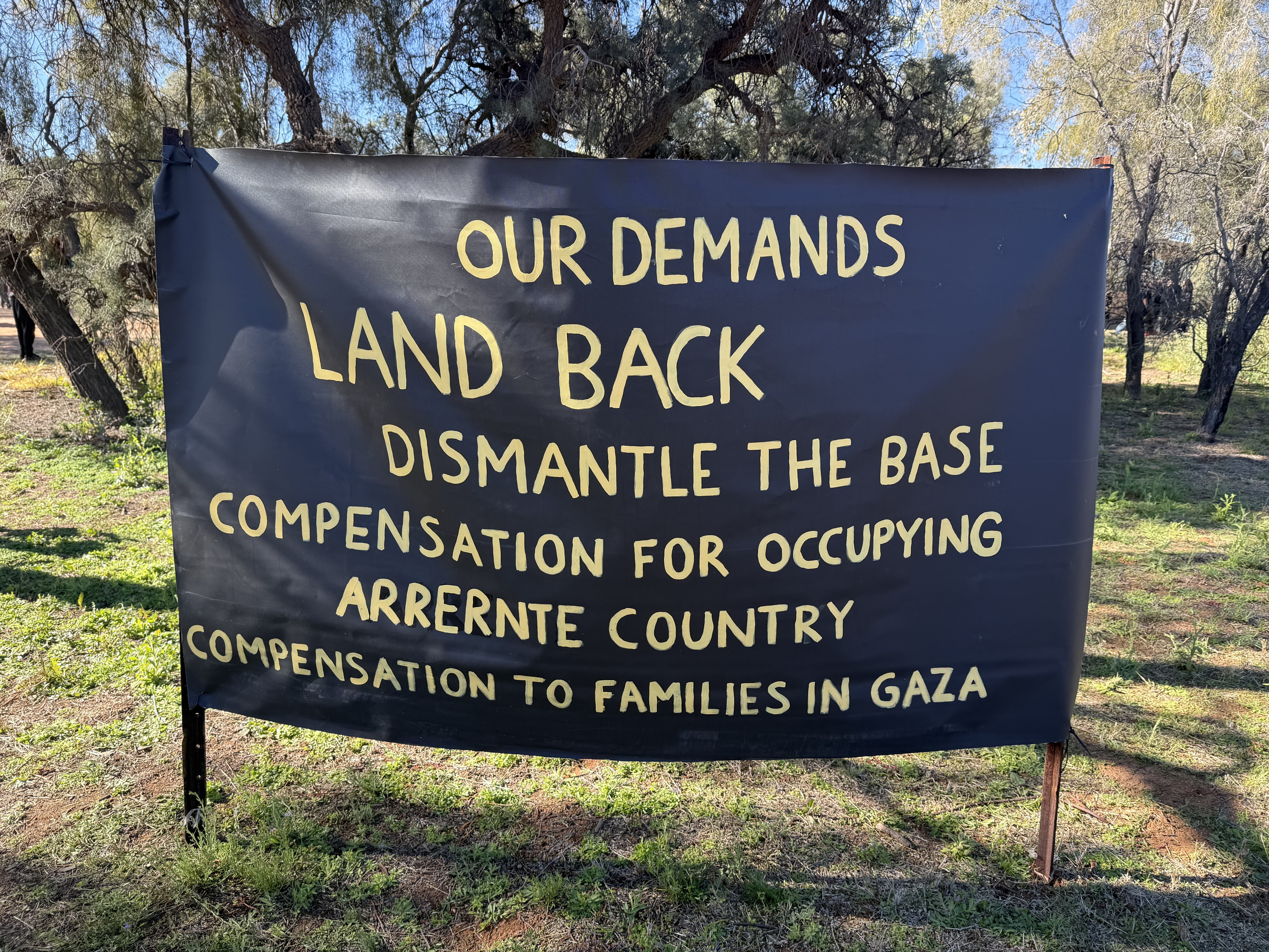 A photo depicting a banner with a list of demands from traditional owners on Arrente Country. There demands are listed as Land Back, dismantly the base, compensation for occupyin Arrernte Country and compansation to families in Gaza.