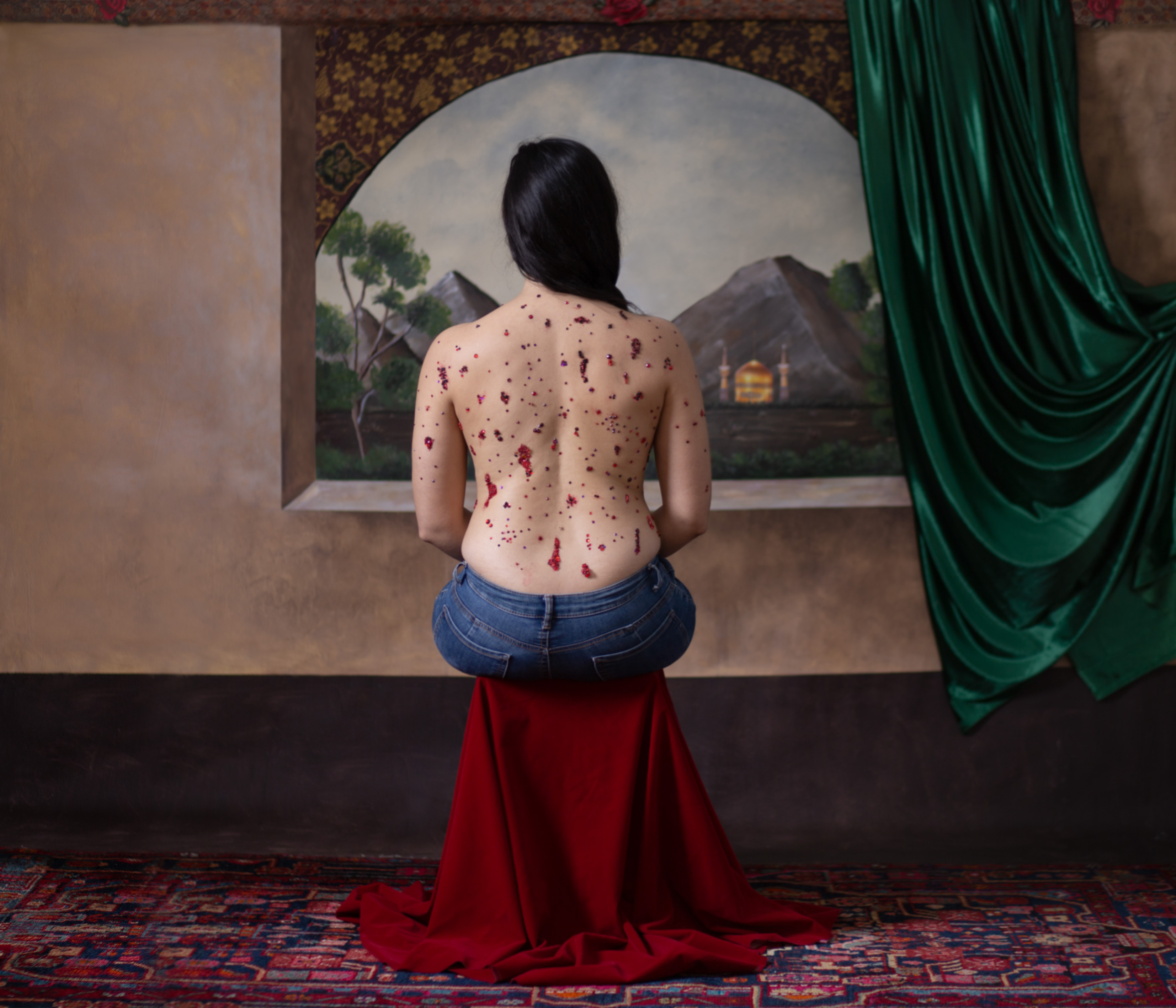 image of a painted background inside a house. There is a woman sitting on on a red stool looking out the window. Her back is faced towards us and is covered in red jewels depicting blood.