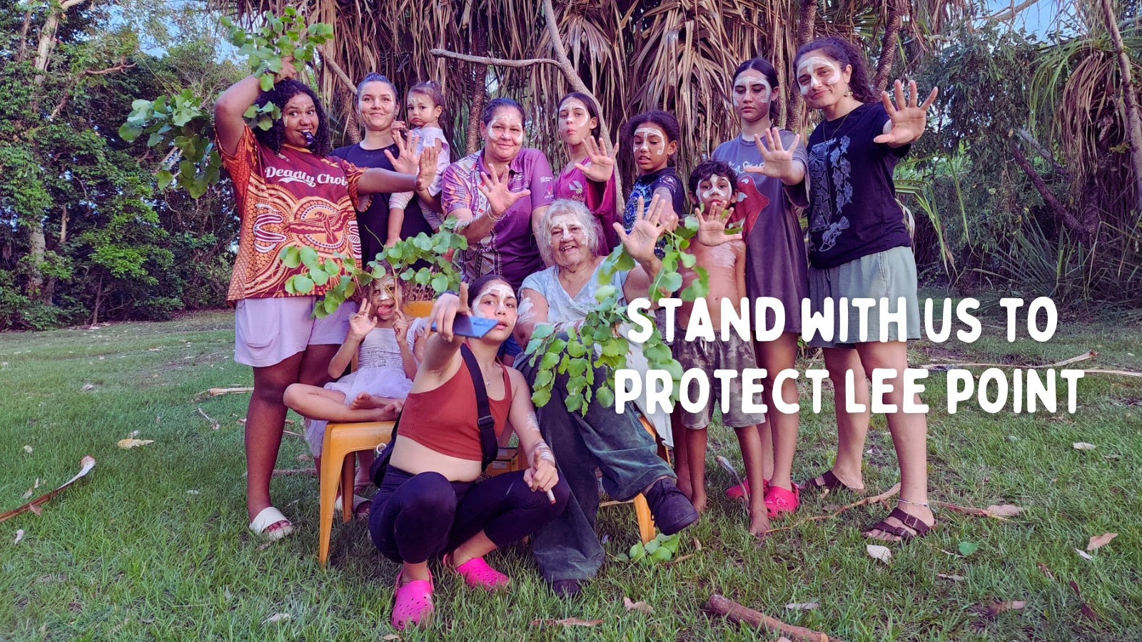A group of Larrakia women and children pose, painted up, in front of lush greenery. Overlaid on the photo is the text "stand with us to save Lee Point."
