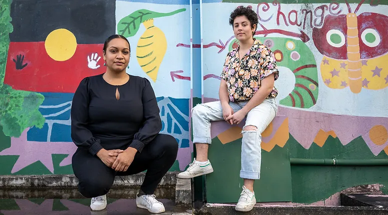 A photograph of Murrawah Johnson and Monique Jeffs in front of a wall painted with a colourful environment-themed mural which includes a large Aboriginal flag. Murrawah is on the left, squatting with hands clasped in front of her, and wears a long-sleeved black shirt, black pants and white sneakers. Monique sits on a protruding feature of the wall on the right, also with their hands clasped in front of them, wearing a floral shirt, torn light blue jeans, and white sneakers. Both have serious expressions.