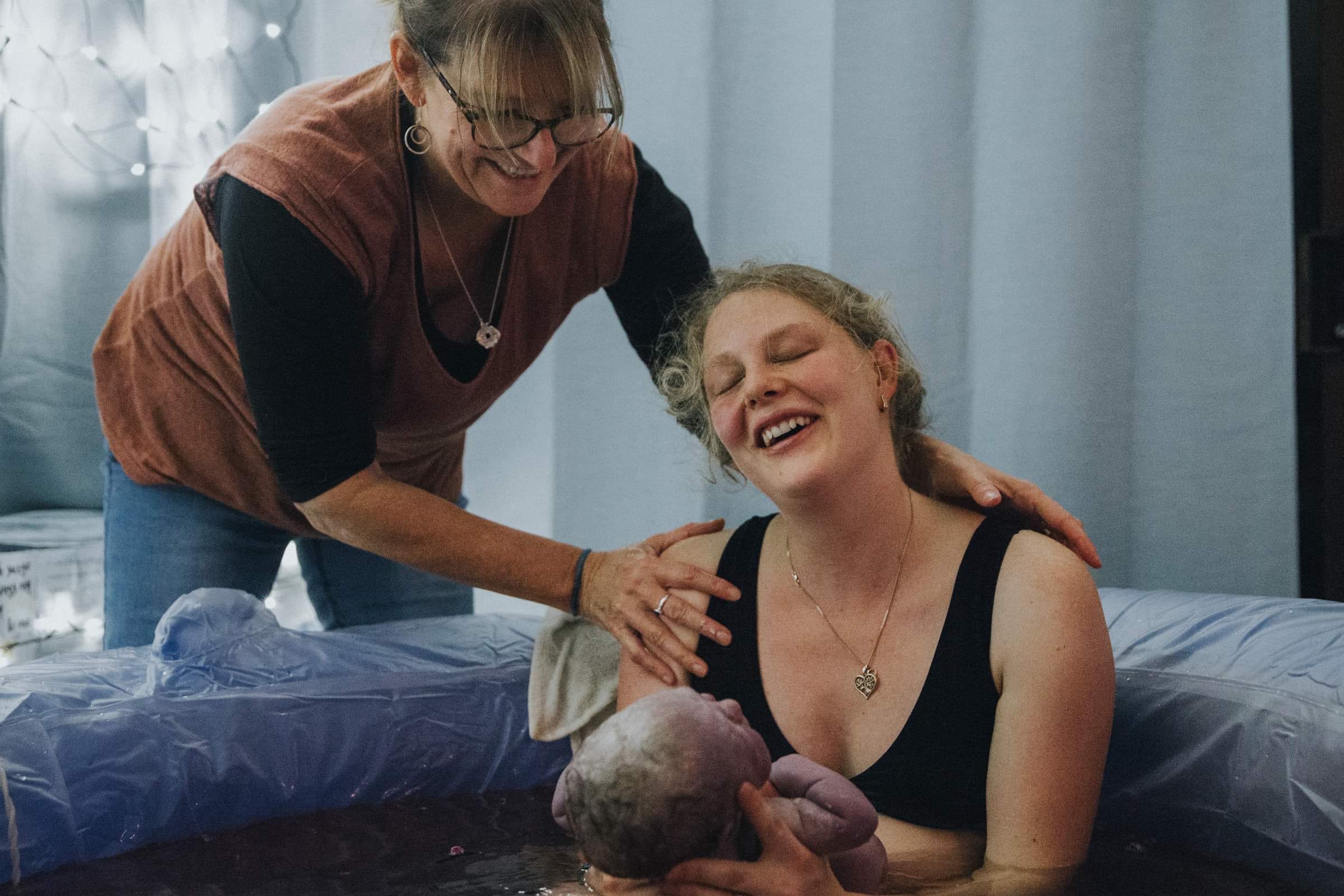 A woman sits in a homebirth pool holding her baby in front of her, she is smiling and has her eyes closed. A woman stand outside of the pool with her hands around her shoulders.