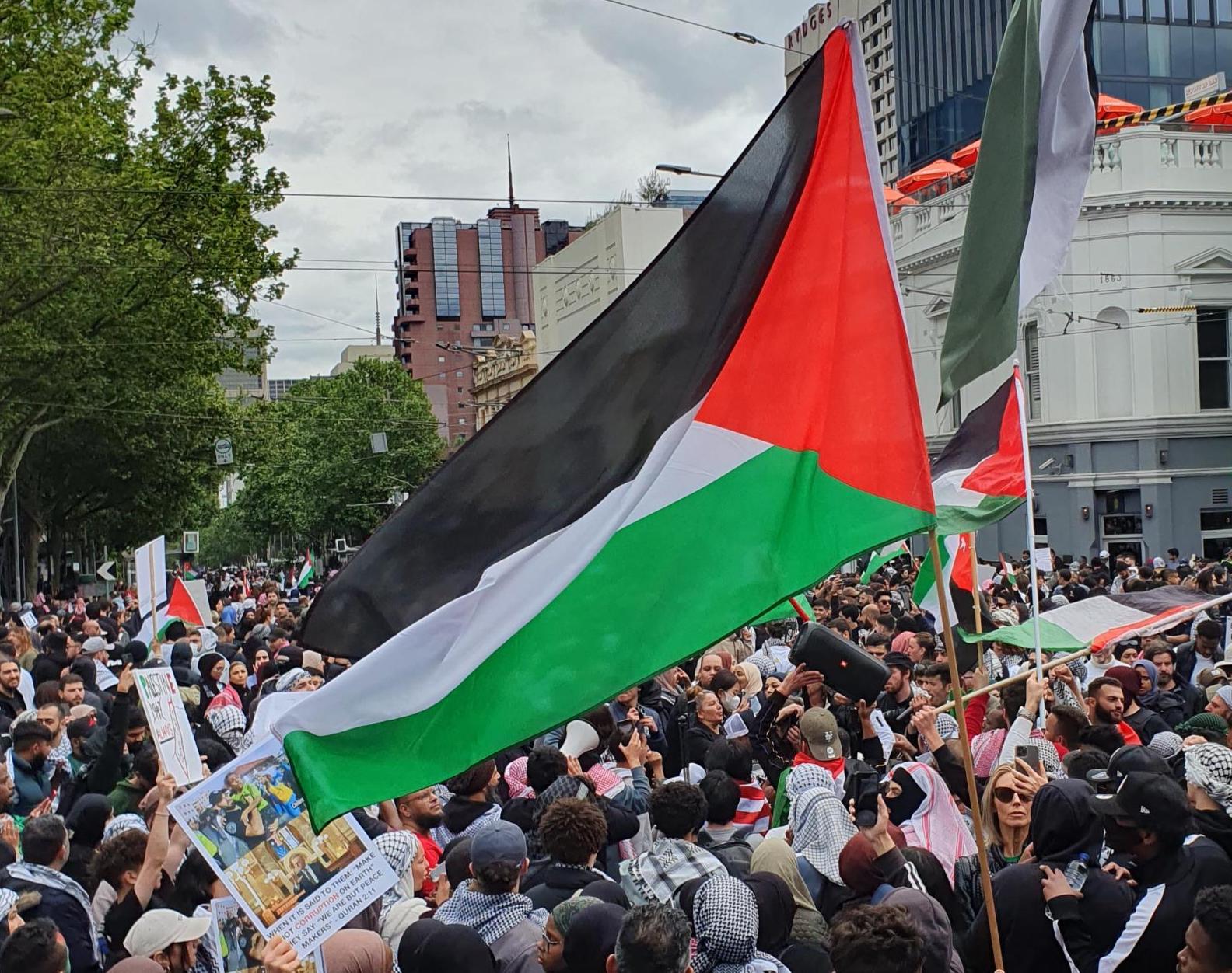 A black, white, red and green Palestinian flag is waved over a large crowd at a rally on Sunday 15th October outside Parliament House in Narrm