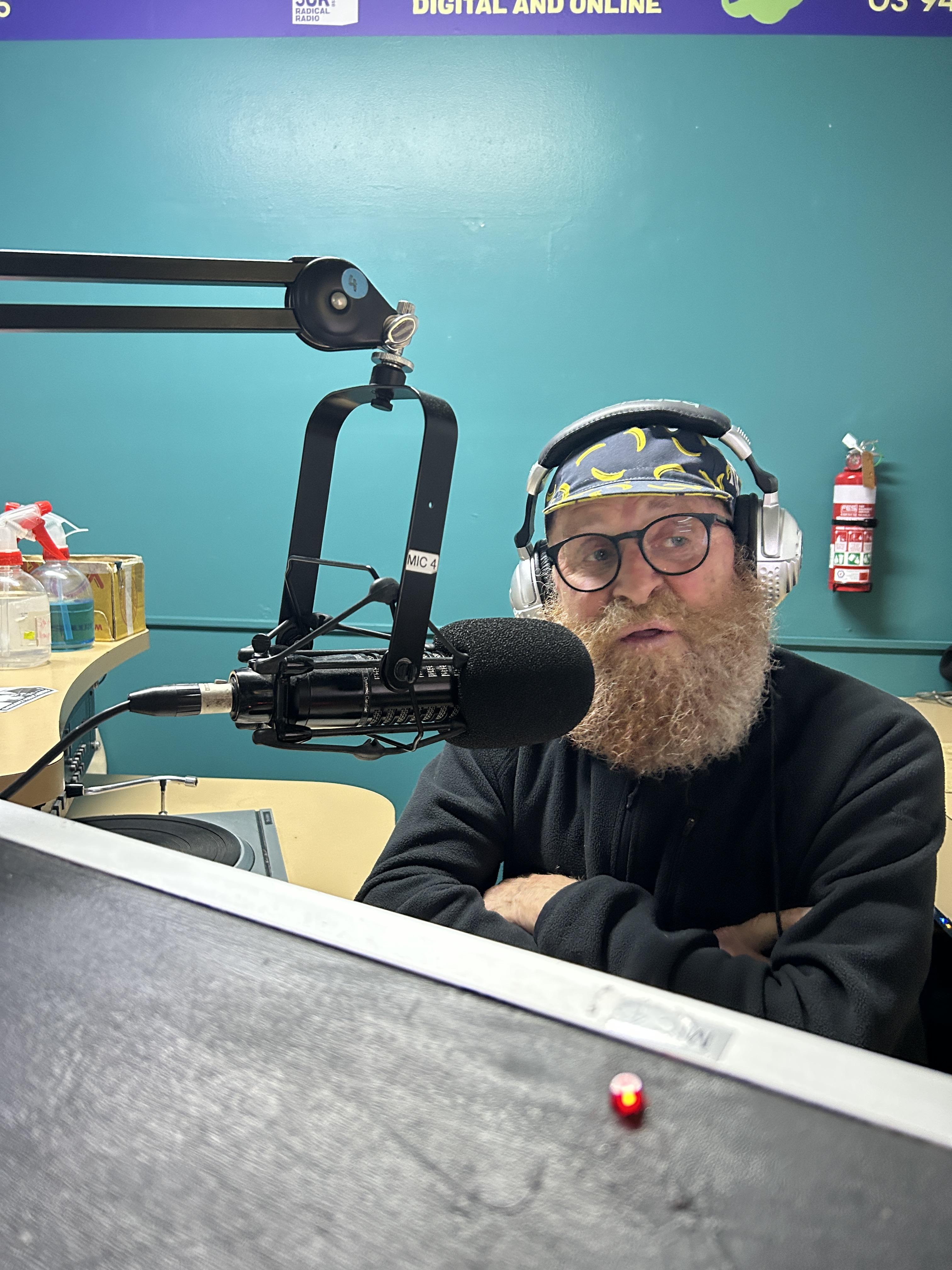 A man with a beard wearing a cycling cap and headphones sits in front of a microphone
