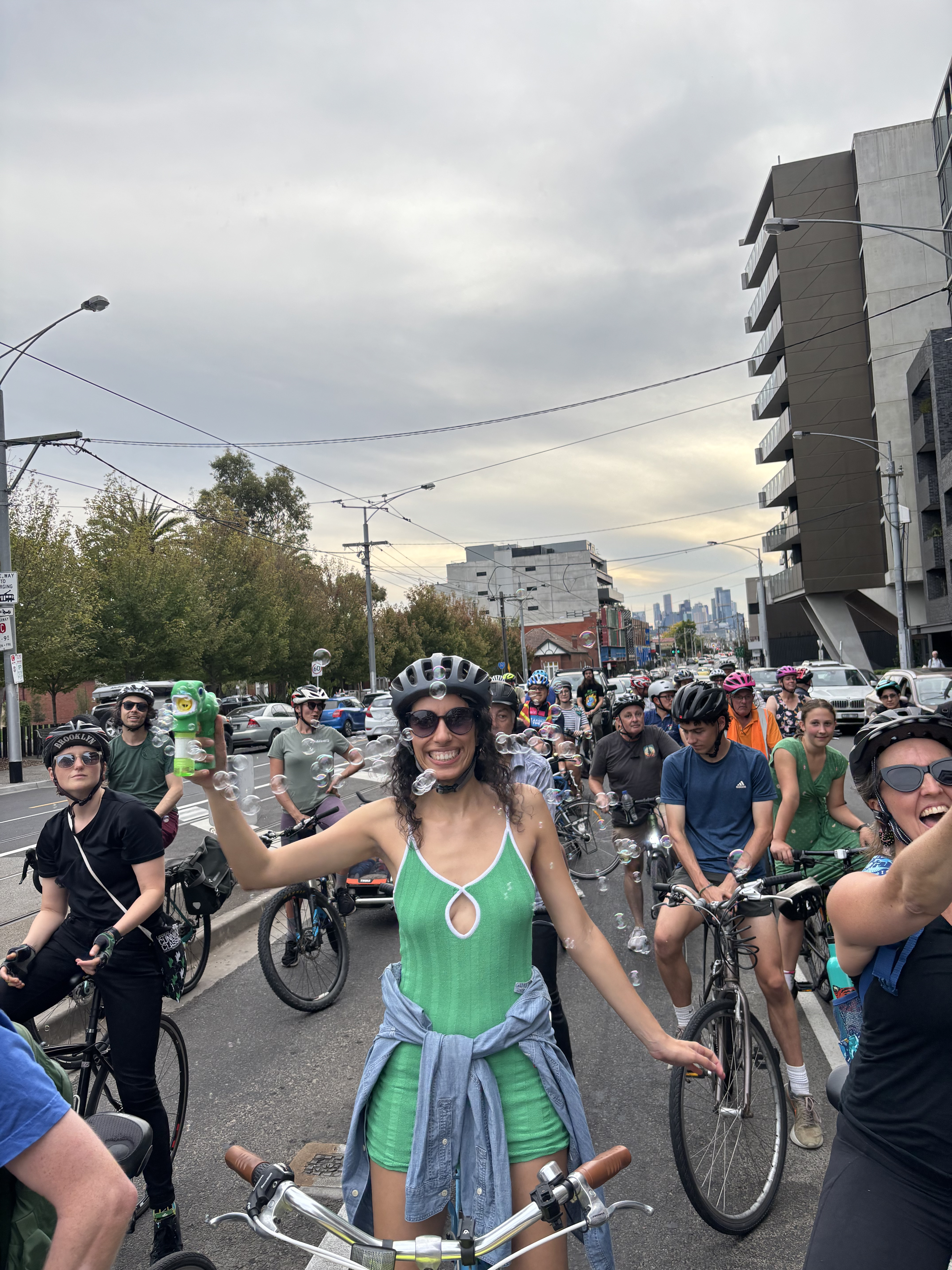 Group of bicycle riders riding on Bridge Road, Richmond, three people smiling at camera, centre person wearing green dress holding a bubble maker and surrounded by bubbles