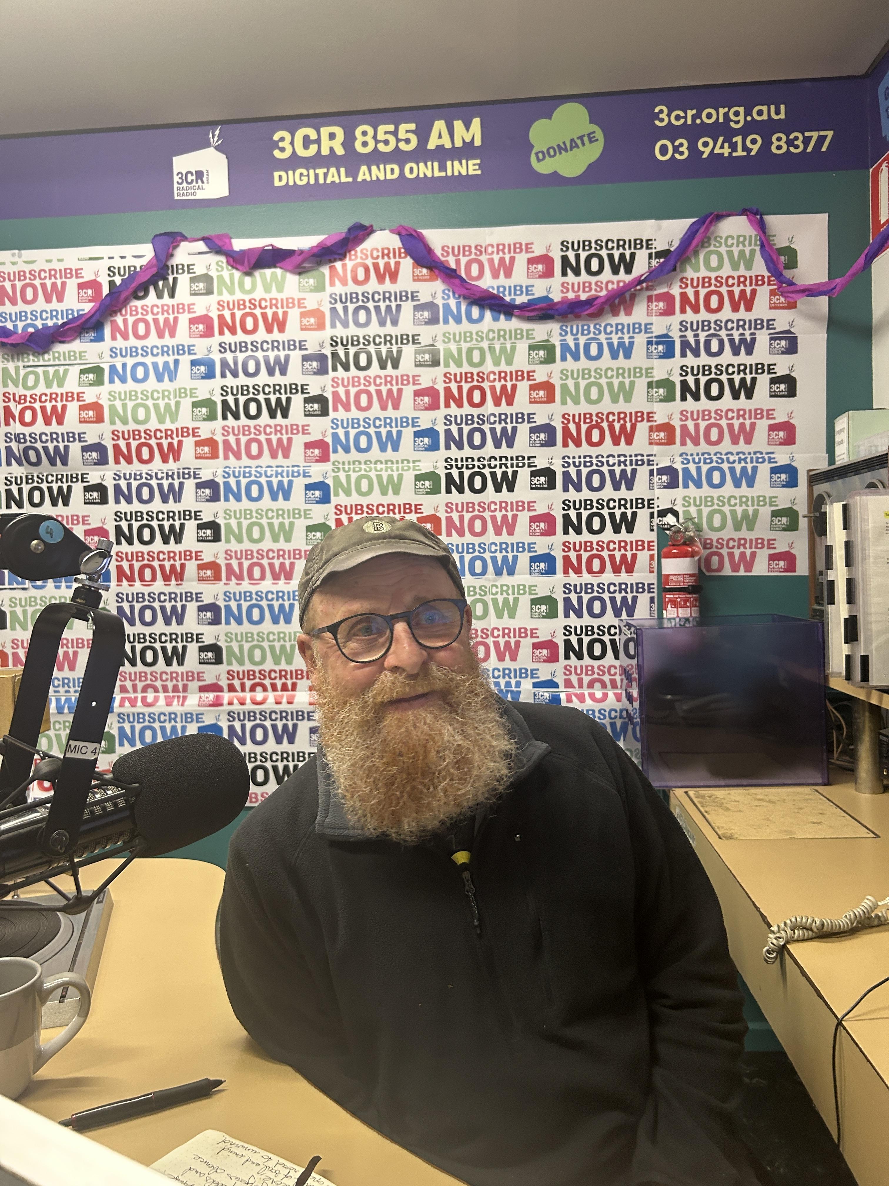 A bearded man sits at the desk with a microphone to one side of him. He is smiling and behind him the wall is covered in Subscribathon material. 