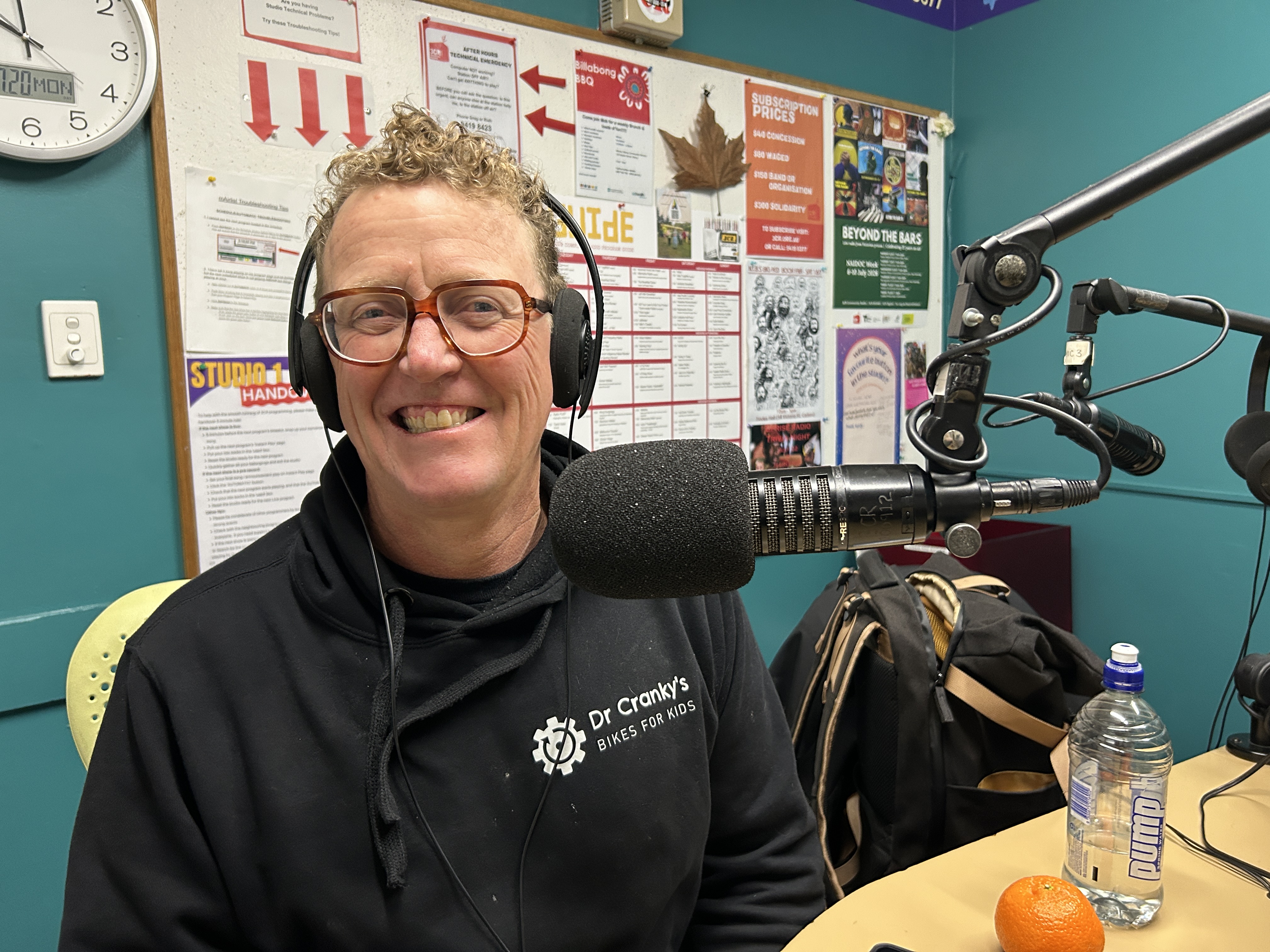 A smiling man wearing headphones sits next to a microphone in the 3CR studio. he is wearing a black sweatshirt with "Dr Cranky's Bikes for kids" written on it. 