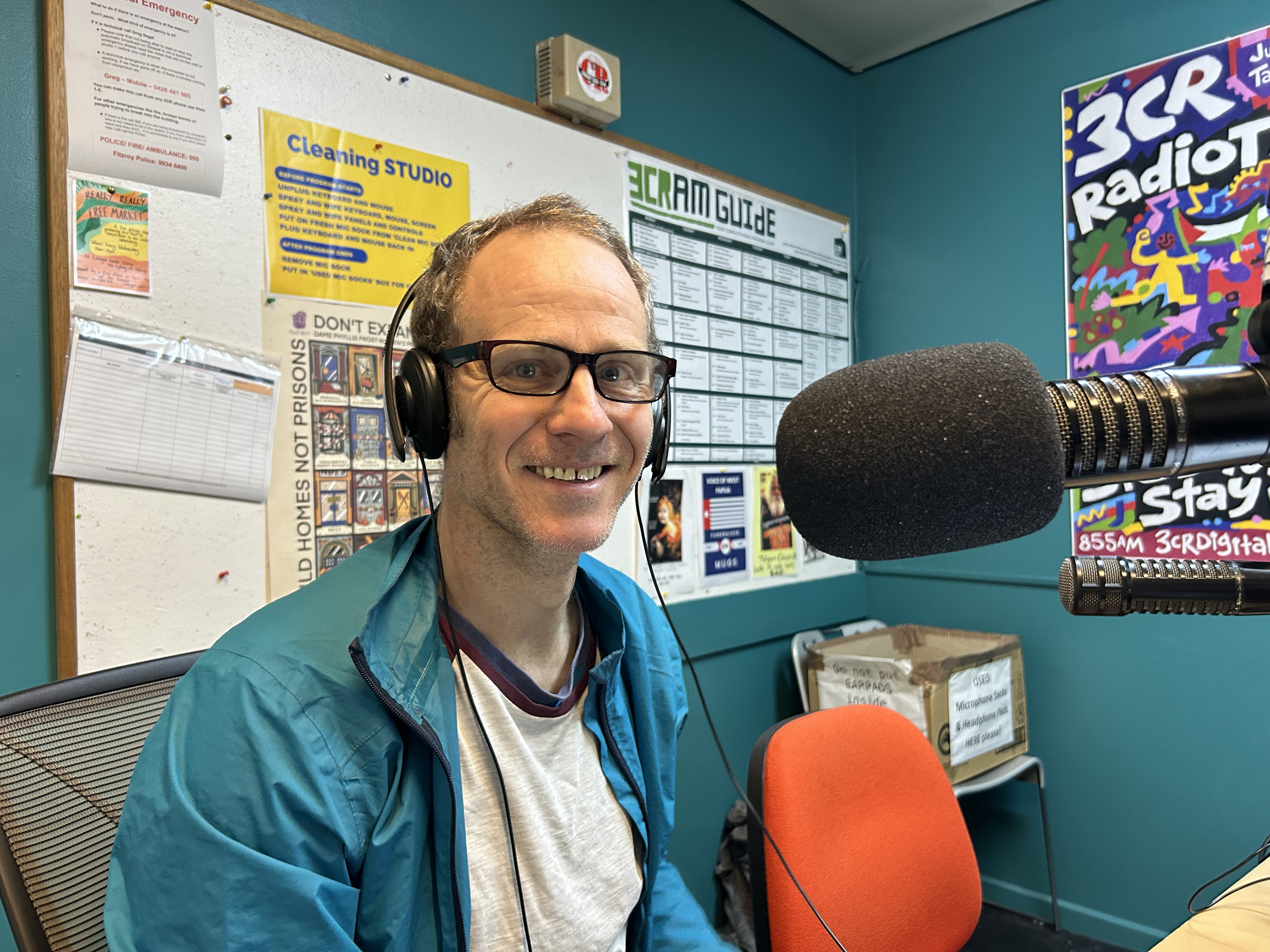 A man sitting facing a microphone in the studios of 3CR. He is looking at the camera and wearing headphones