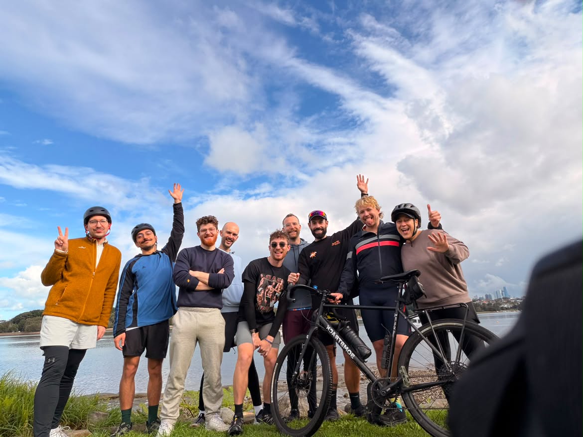 Group of casually attired bicycle riders smiles at the camera, clouds and blue sky behind them.