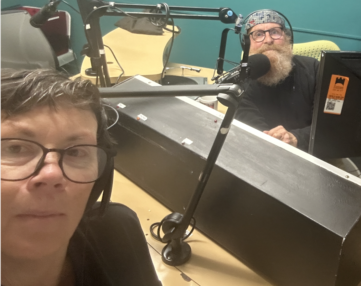 A woman wearing glasses looks into the camera, nehind her is a panelling dek and behind that a bearded man looking at the camera. 