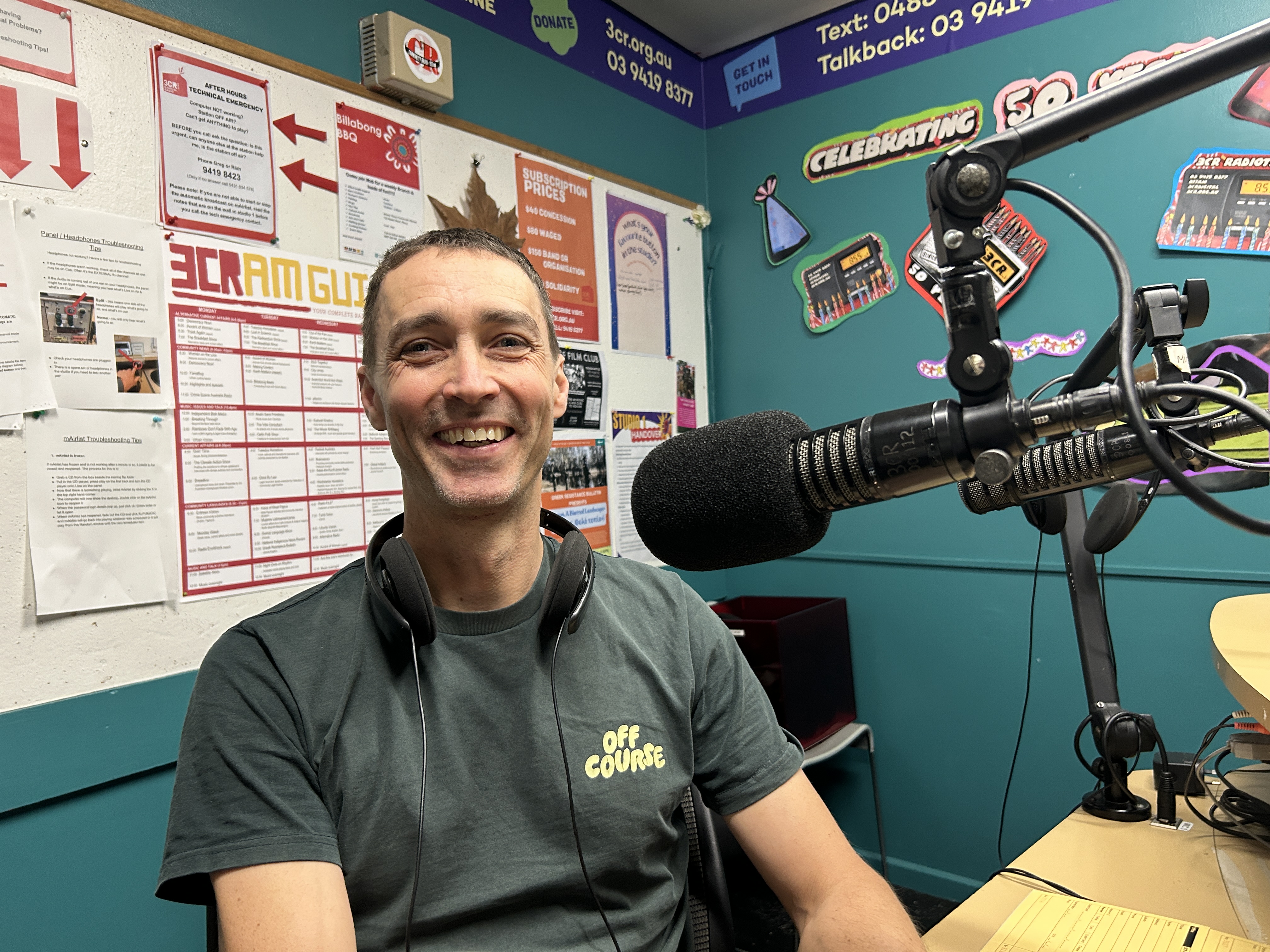 A smiling man in a green t-shirt with the logo "Off Course" on it sits next to a microphone and looks at the camera. 