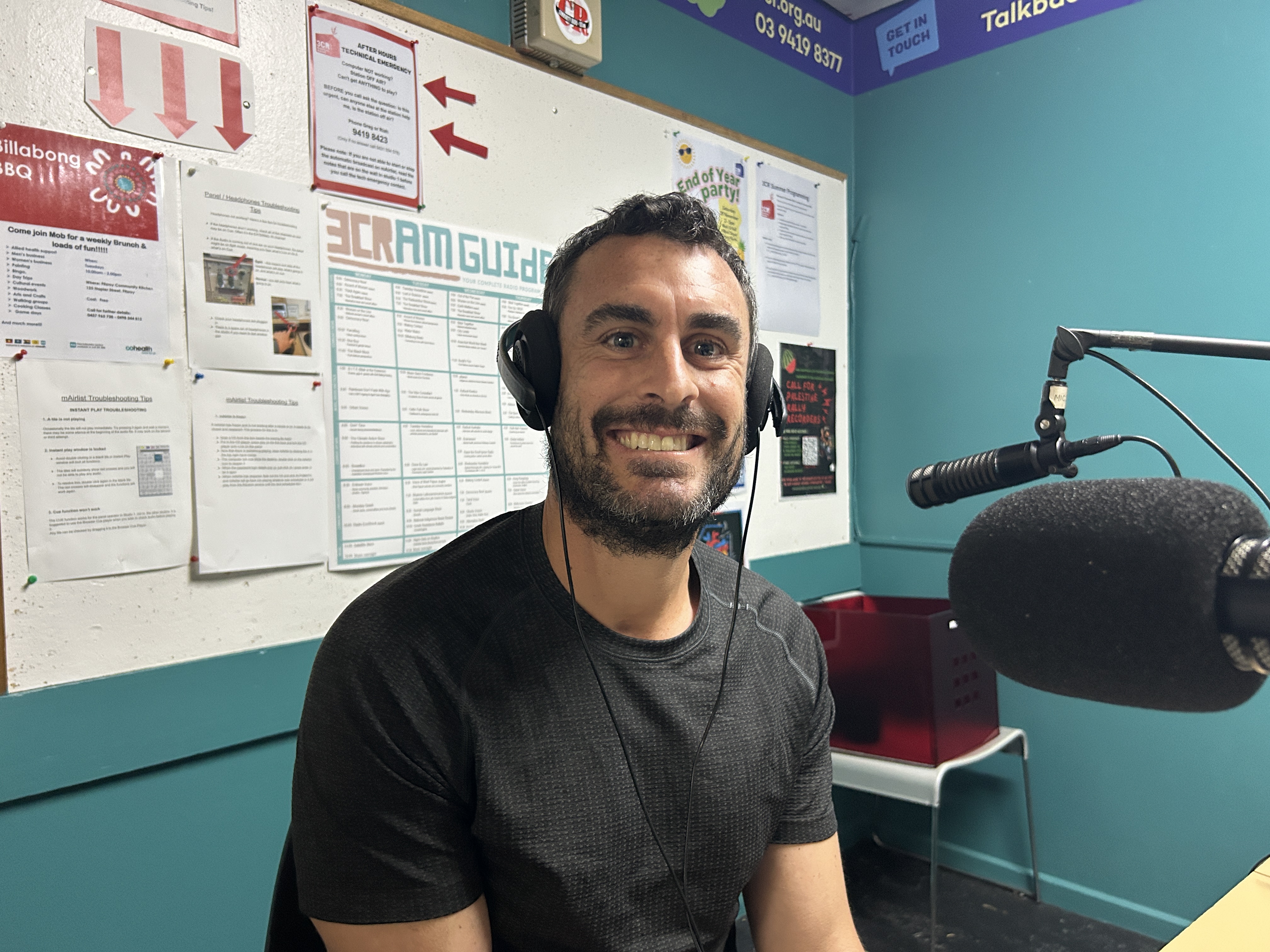 A man in a black t-shirt sits smailing at the camera. he has headphones on his head and is sitting in front of a microphone