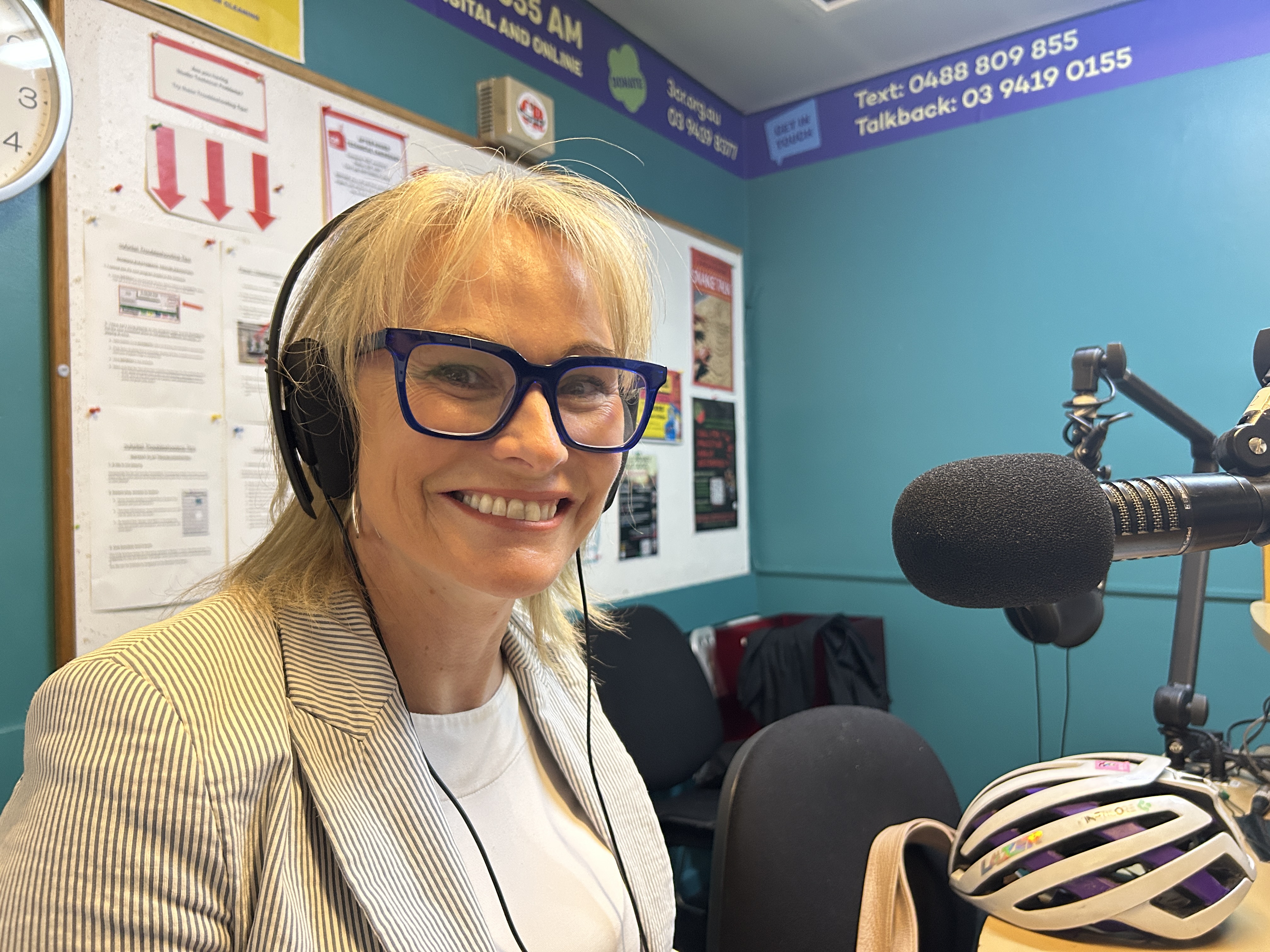 A blonde woman smiles at the camera while wearing headphones. She is sitting in front of a microphone. 
