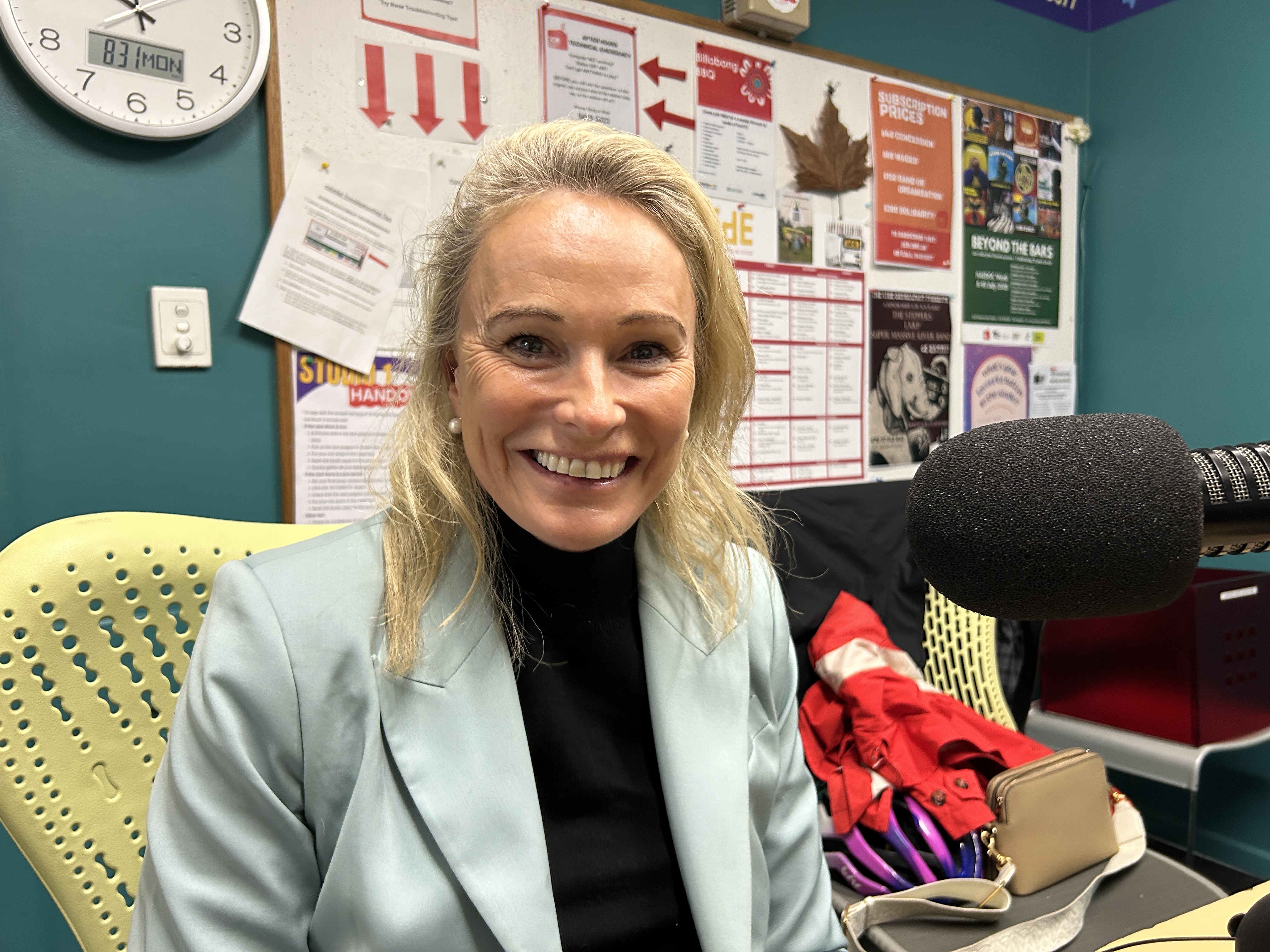 A smiling woman with blonde hair in a pale blue blazer sits in the studio at 3CR looking at the camera. 