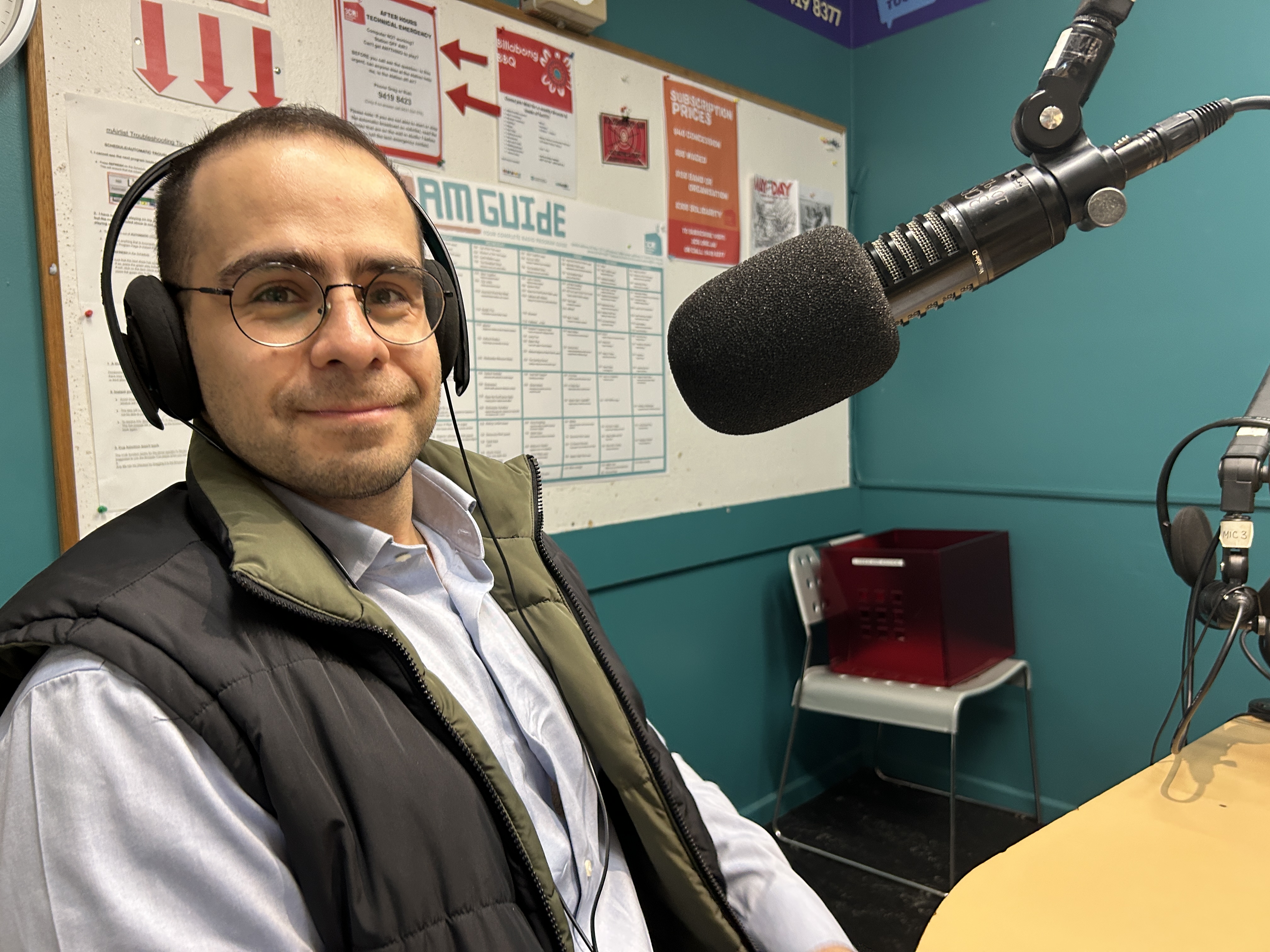 A man wearing headphones sits in front of a microphone and faces the camera