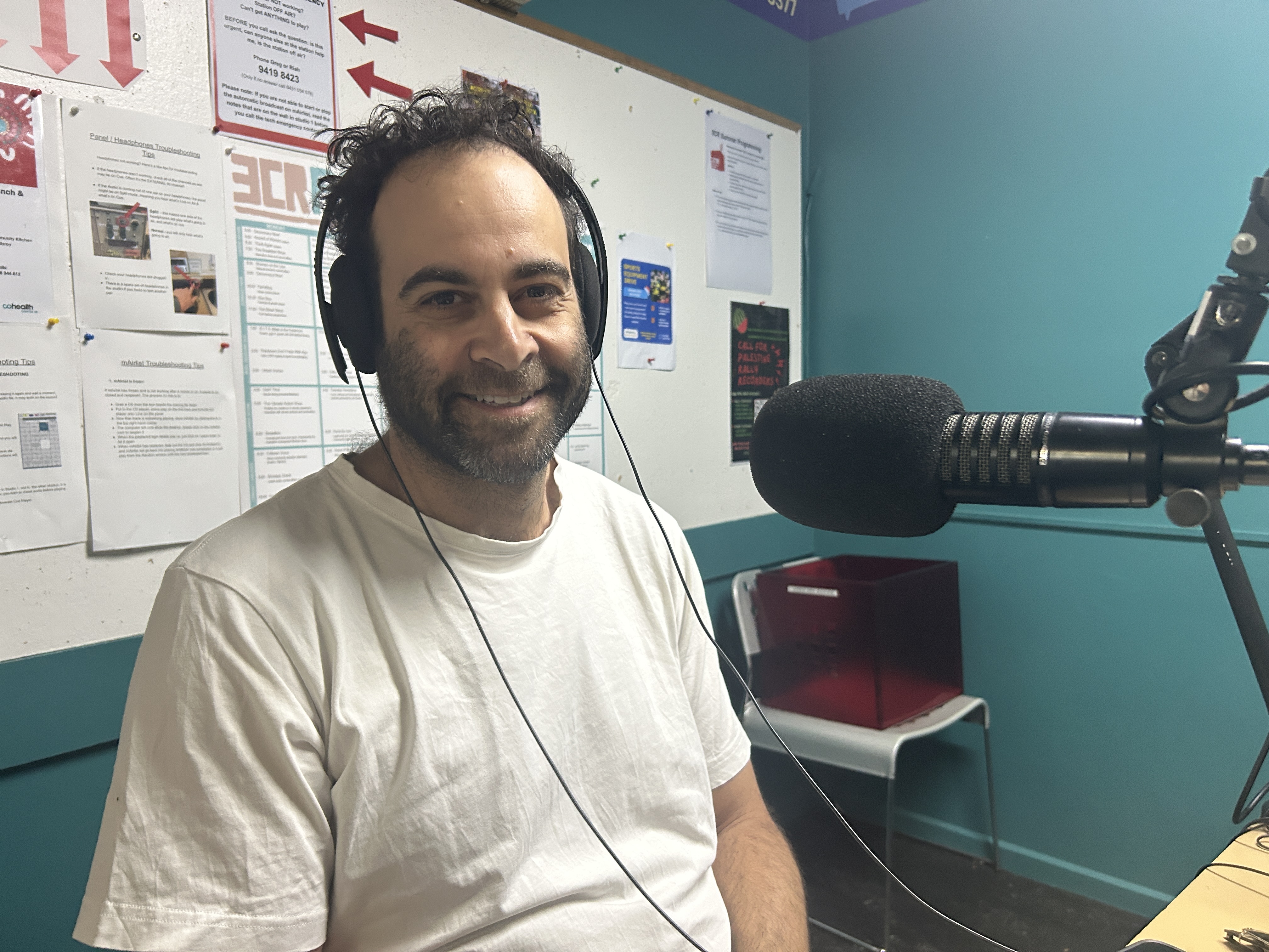 A bearded man in a white t-shirt siots in the 3CR studio. He has a microphone in front of him and is smiling at the camera. 