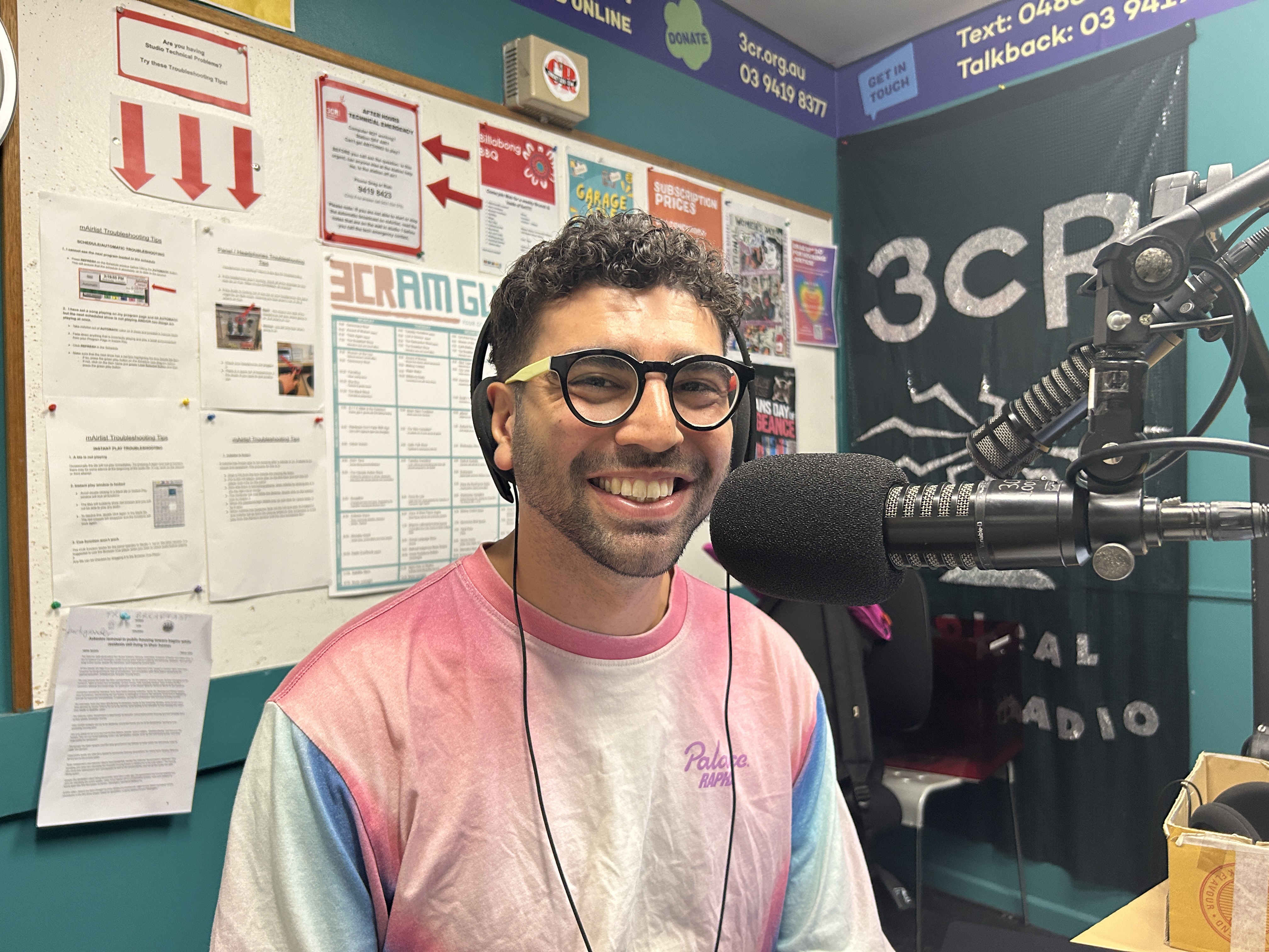 A smiling man wearing glasses and a dope longsleeved tee sits in front of a microphone in the studio at 3CR