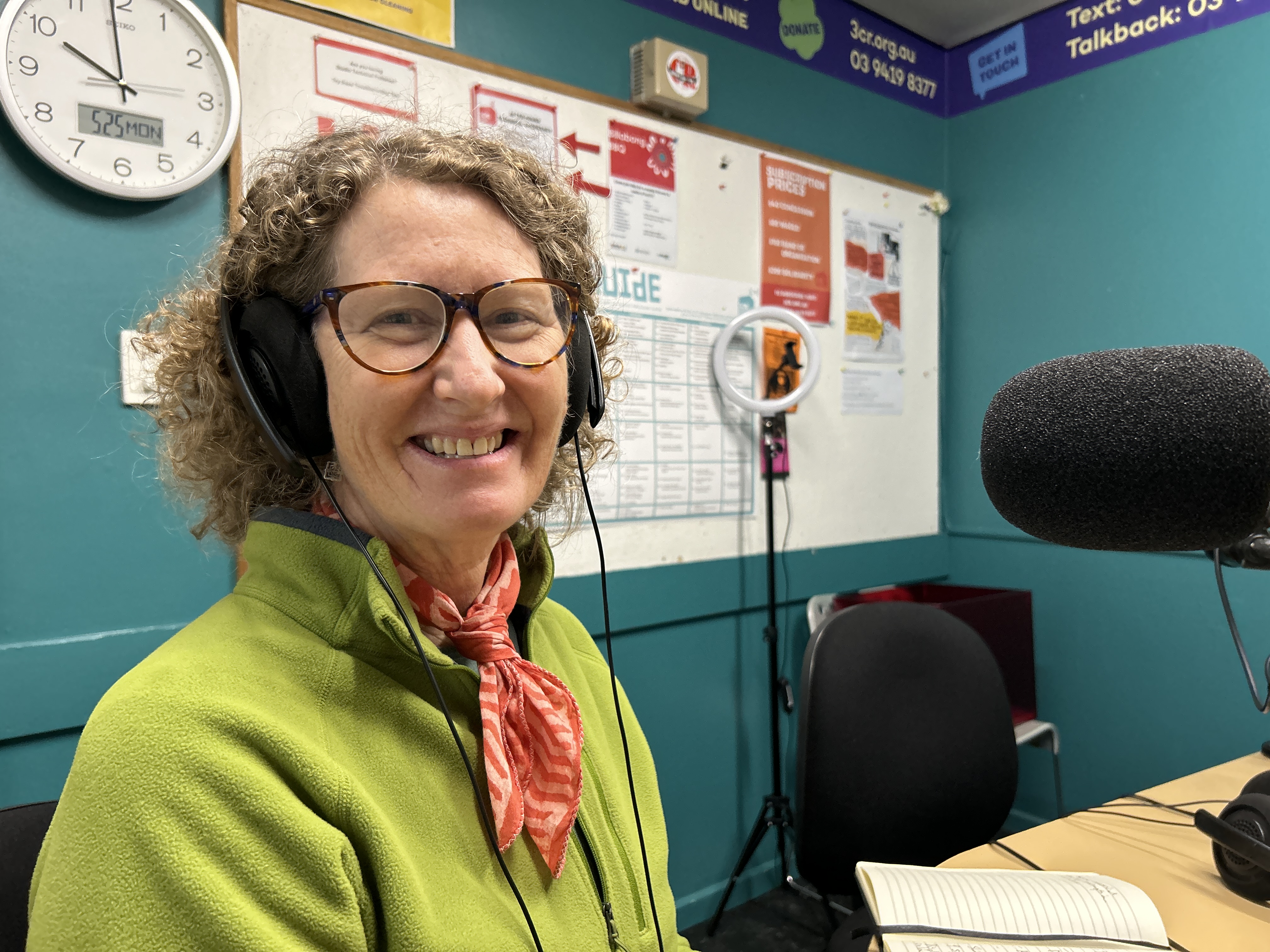 A smiling woman with curly hair sits in front of a microphone. She is wearing glasses, a green top and headphones. 