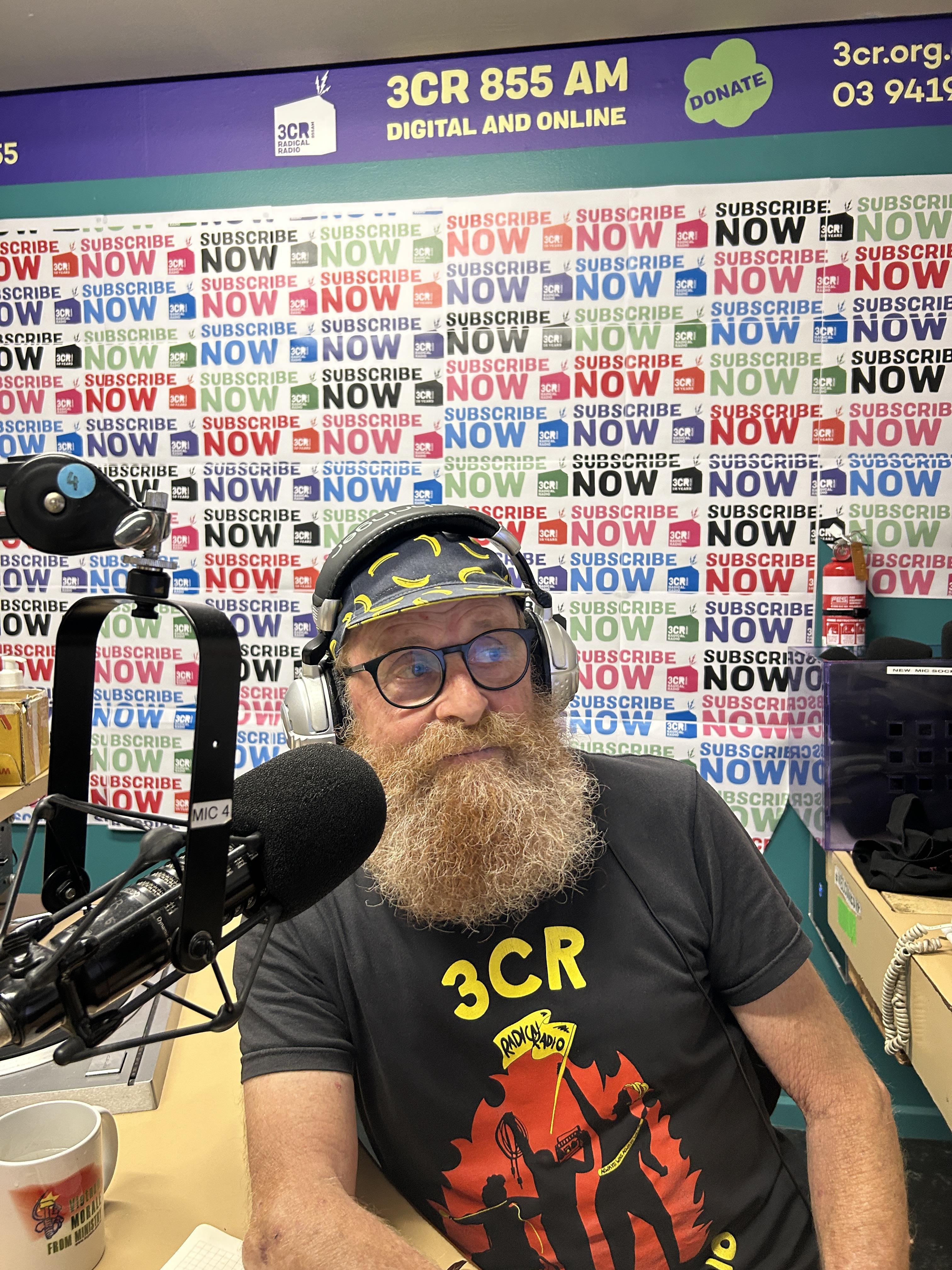 A bearded man wearing headphones sits in front of a microphone. behind him is wall poster with Subscribe on it repeatedly. 