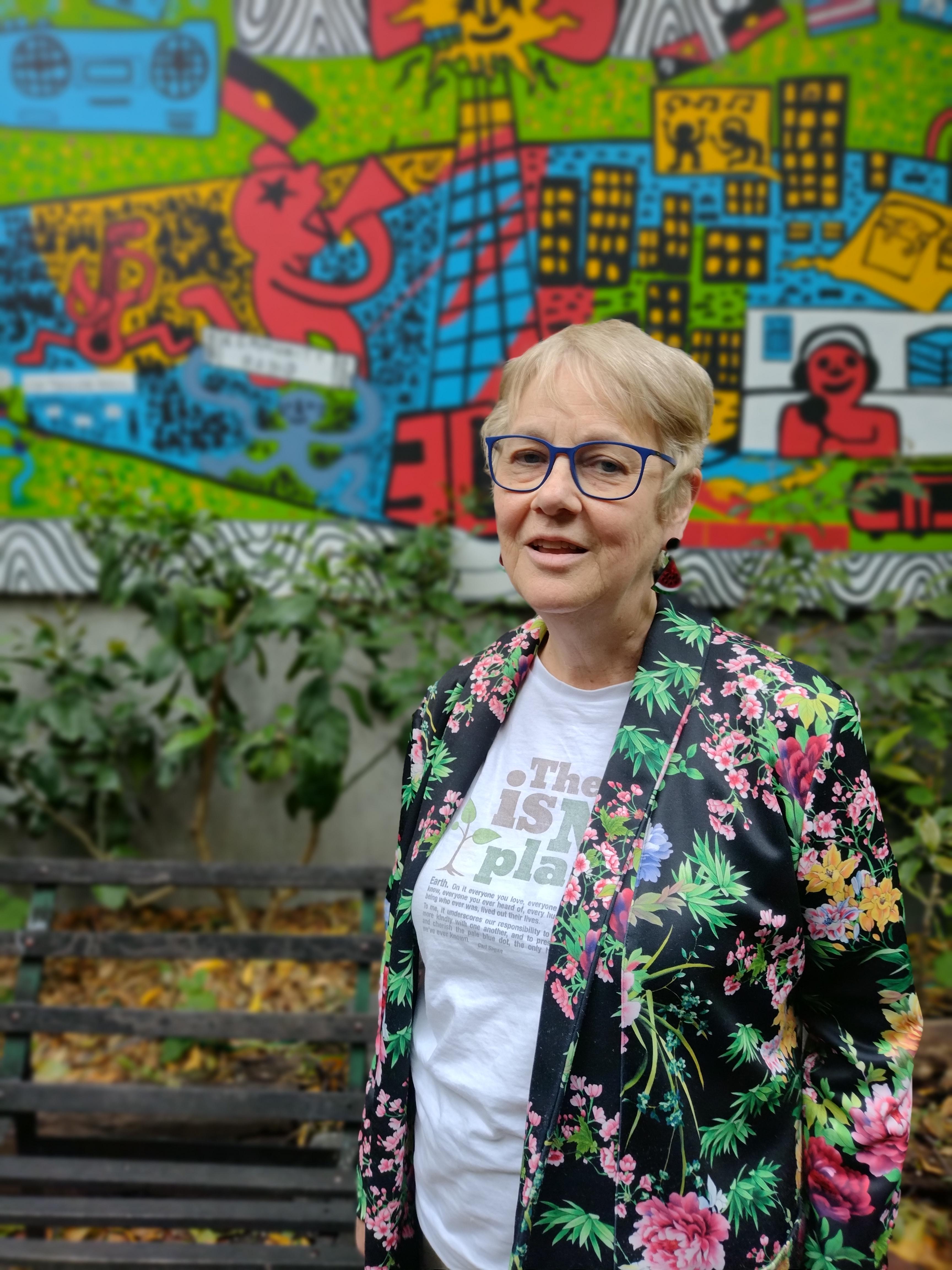 An older woman wearing a colourful jacket and short hair looks at the camera, she's standing outside in front of a mural and she looks content