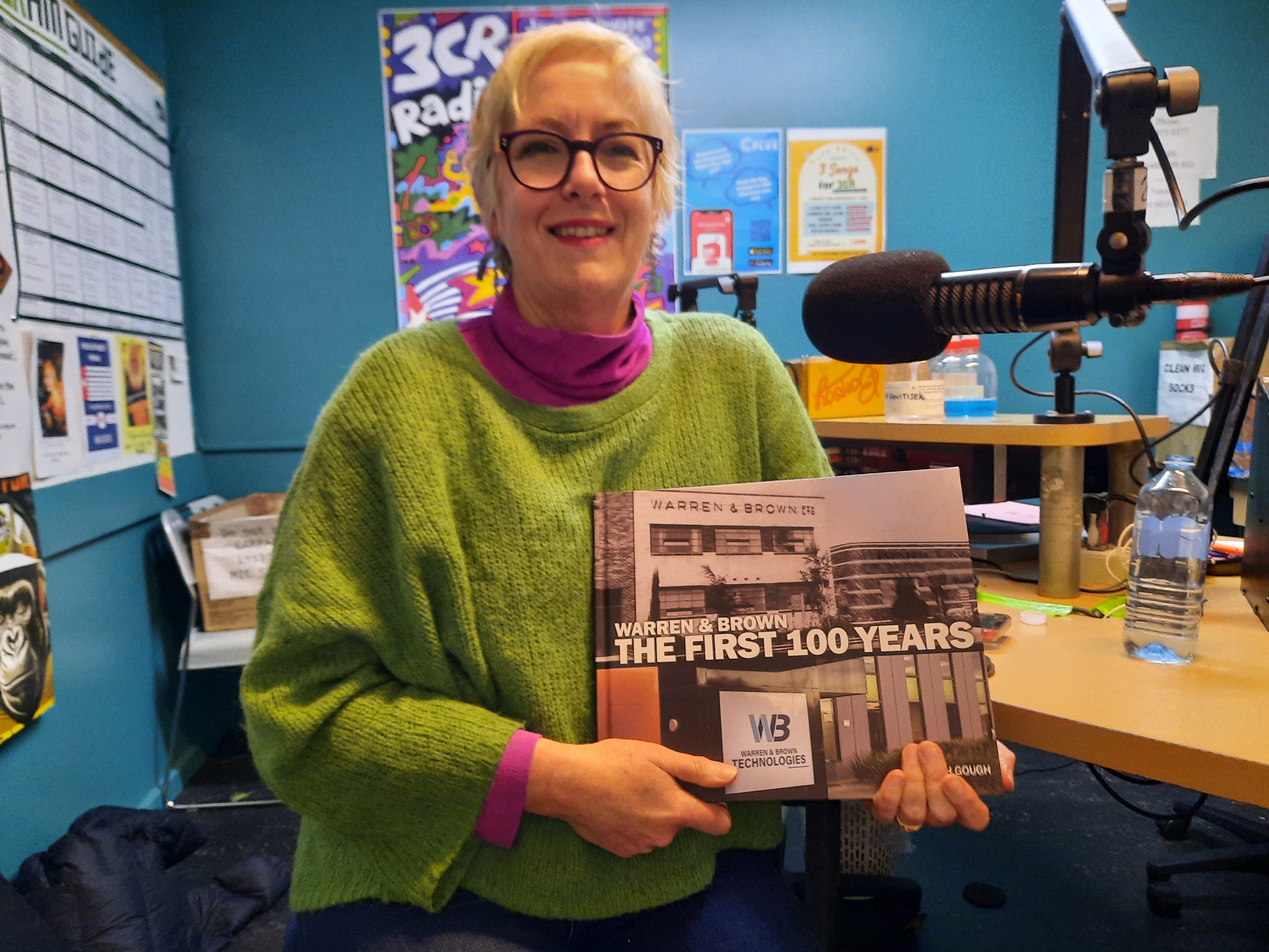 Deborah sits in studio 1 at 3CR holding her book and smiling at the viewer. She wears a green and pink jumper and sports black glasses