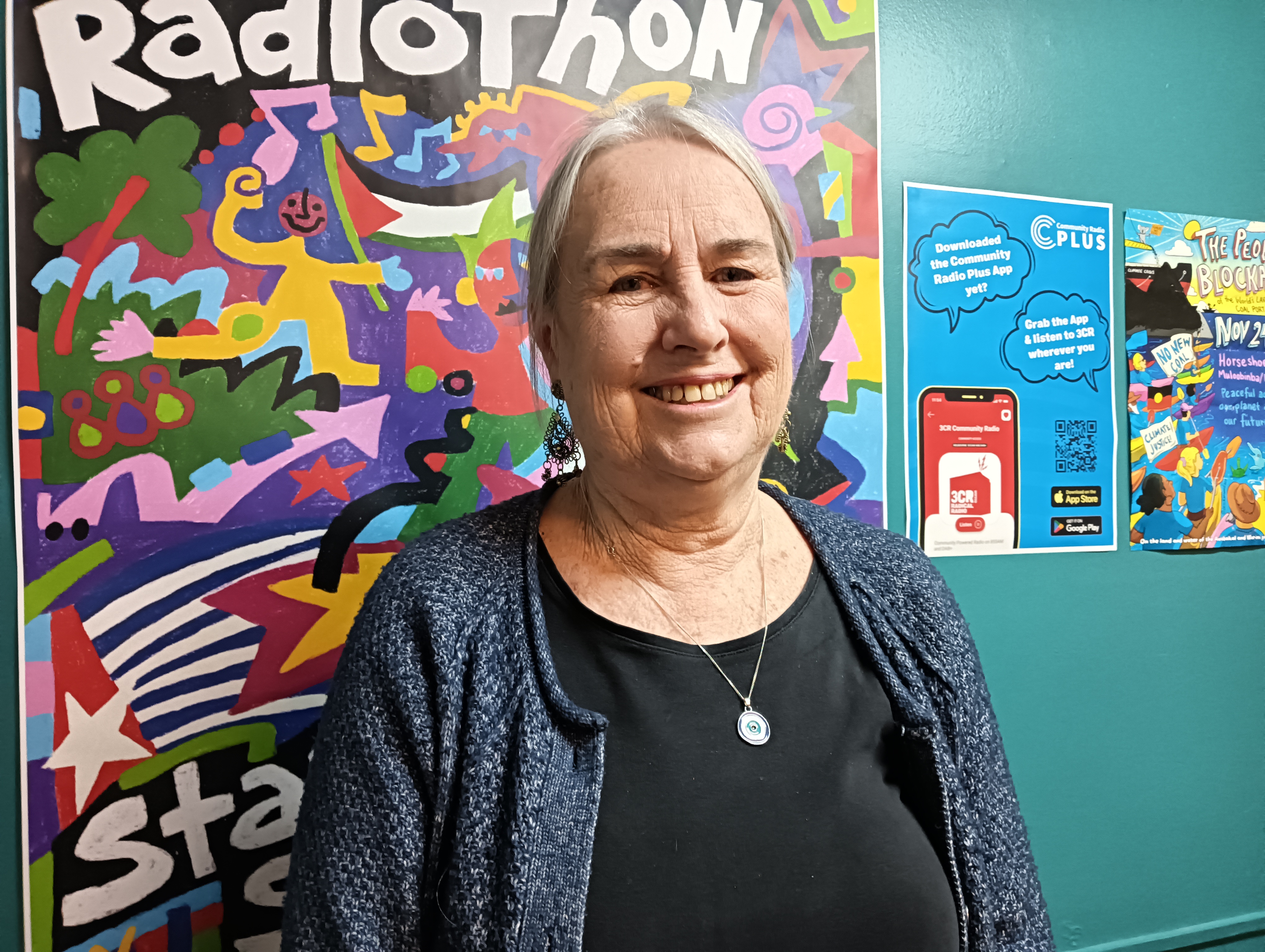 Sue looks towards the viewer wearing a black dress and blue cardigan. She stands in front of this year's colourful 3CR Radiothon poster and smiles 