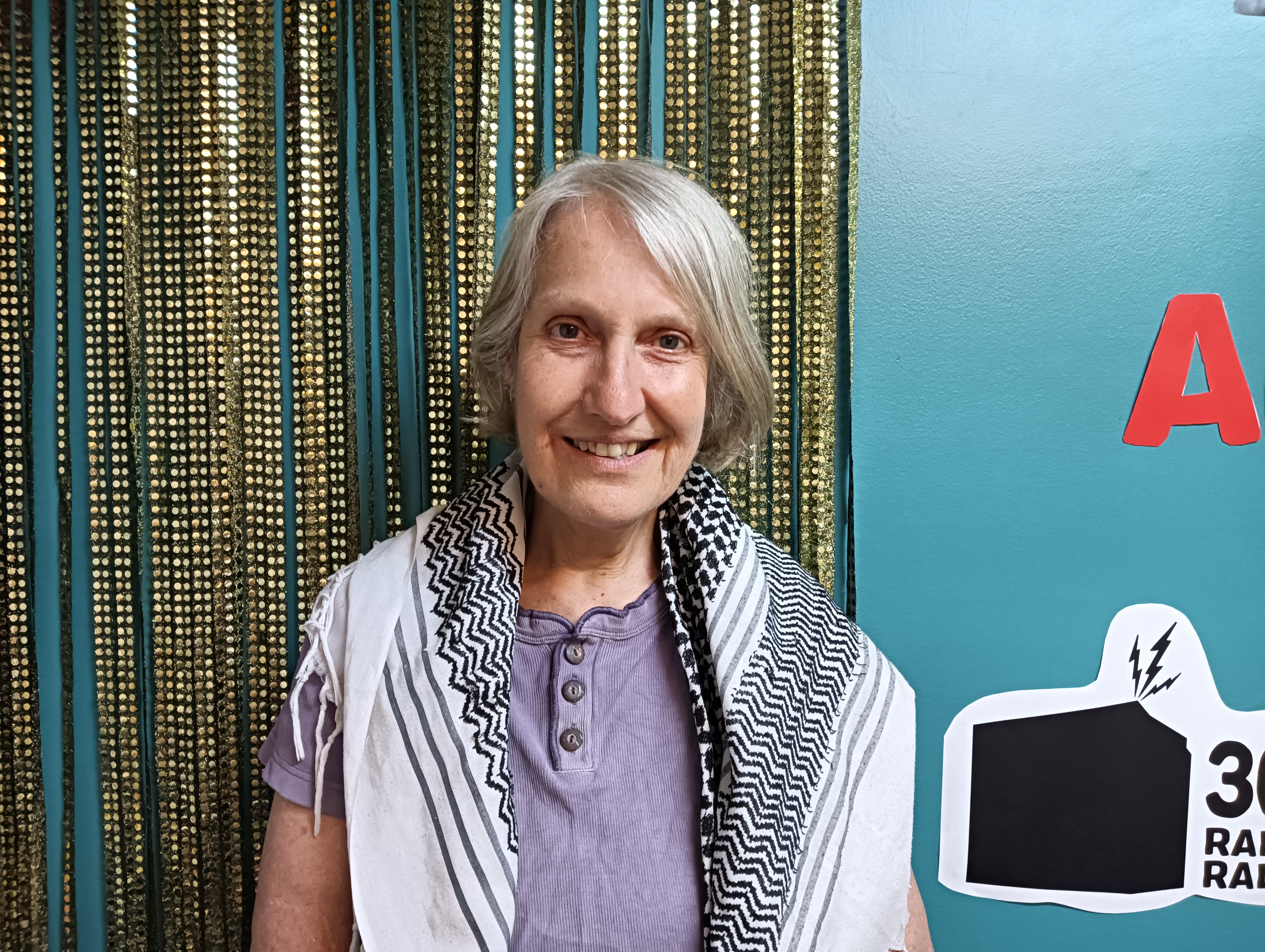Sue stands in studio one at 3CR. Behind her is a gold sequined curtain. Sue wears a Palestinian kufiya around her shoulders.
