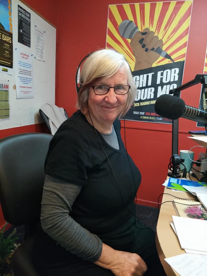 A woman with grey hair is looking at the camera, she is in a radio studio wearing headphones, glasses and a black shirt with a grey undershirt. 