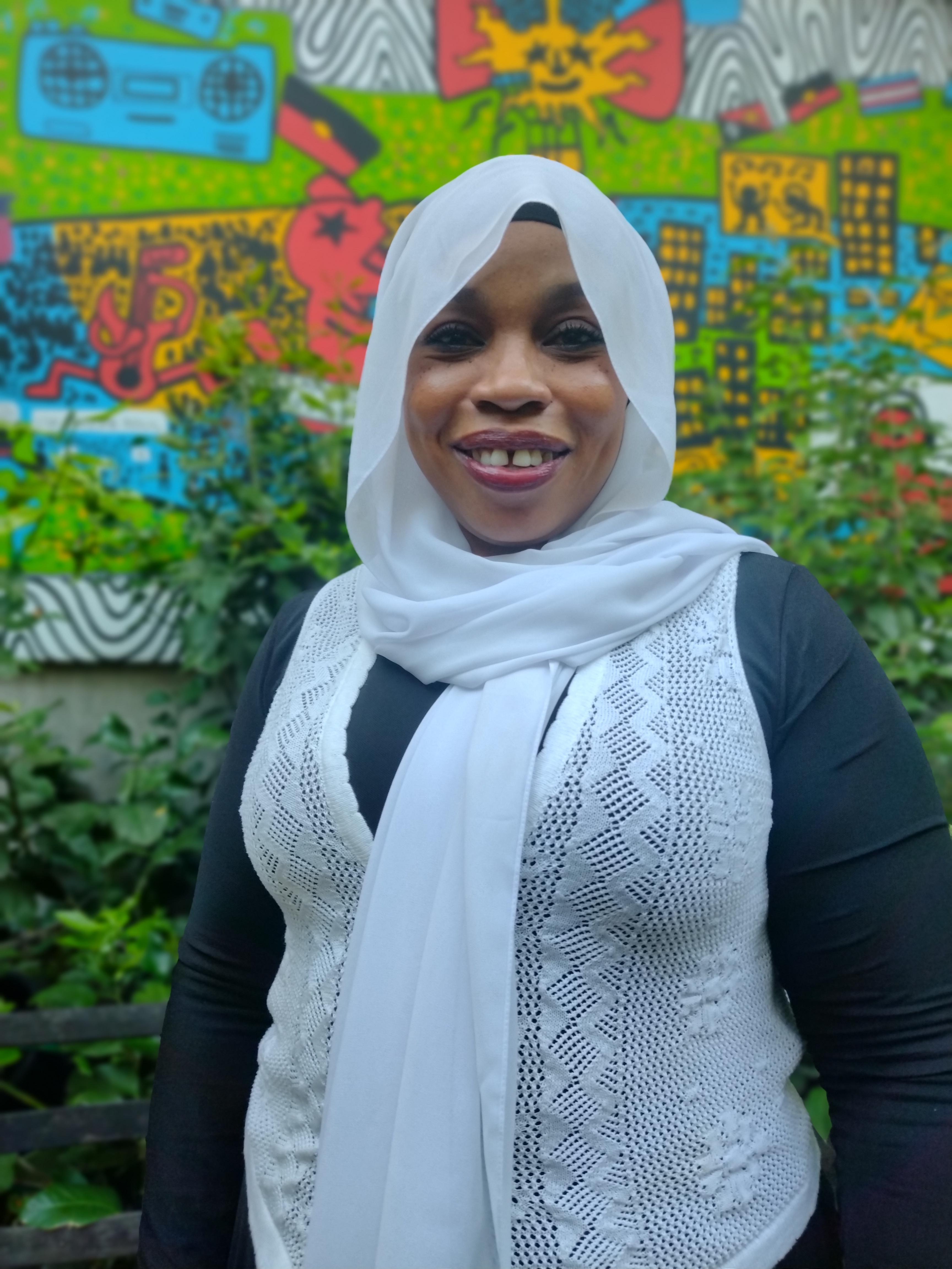 An image of a muslim woman, she is wearing a white hijab and has a beautiful smile. She is standing in front of a colourful mural in a courtyard. 