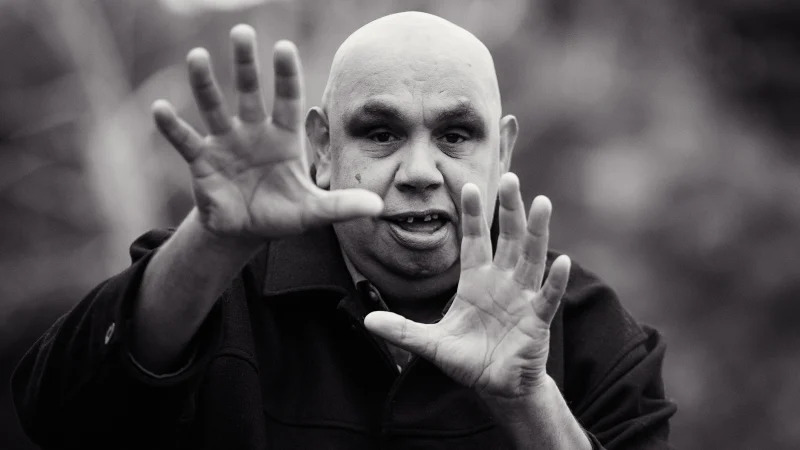 A black-and-white image of Kutcha with his arms raised in front of him making an expressive hand gesture towards the viewer