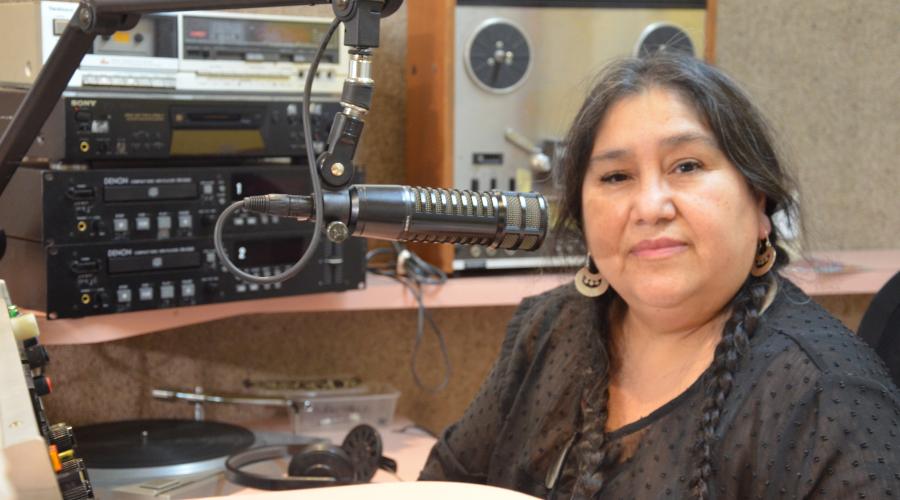 A woman sits in a radio studio among equipment. She wears a black top and has long black hair. She looks with a gentle smile towards the viewer